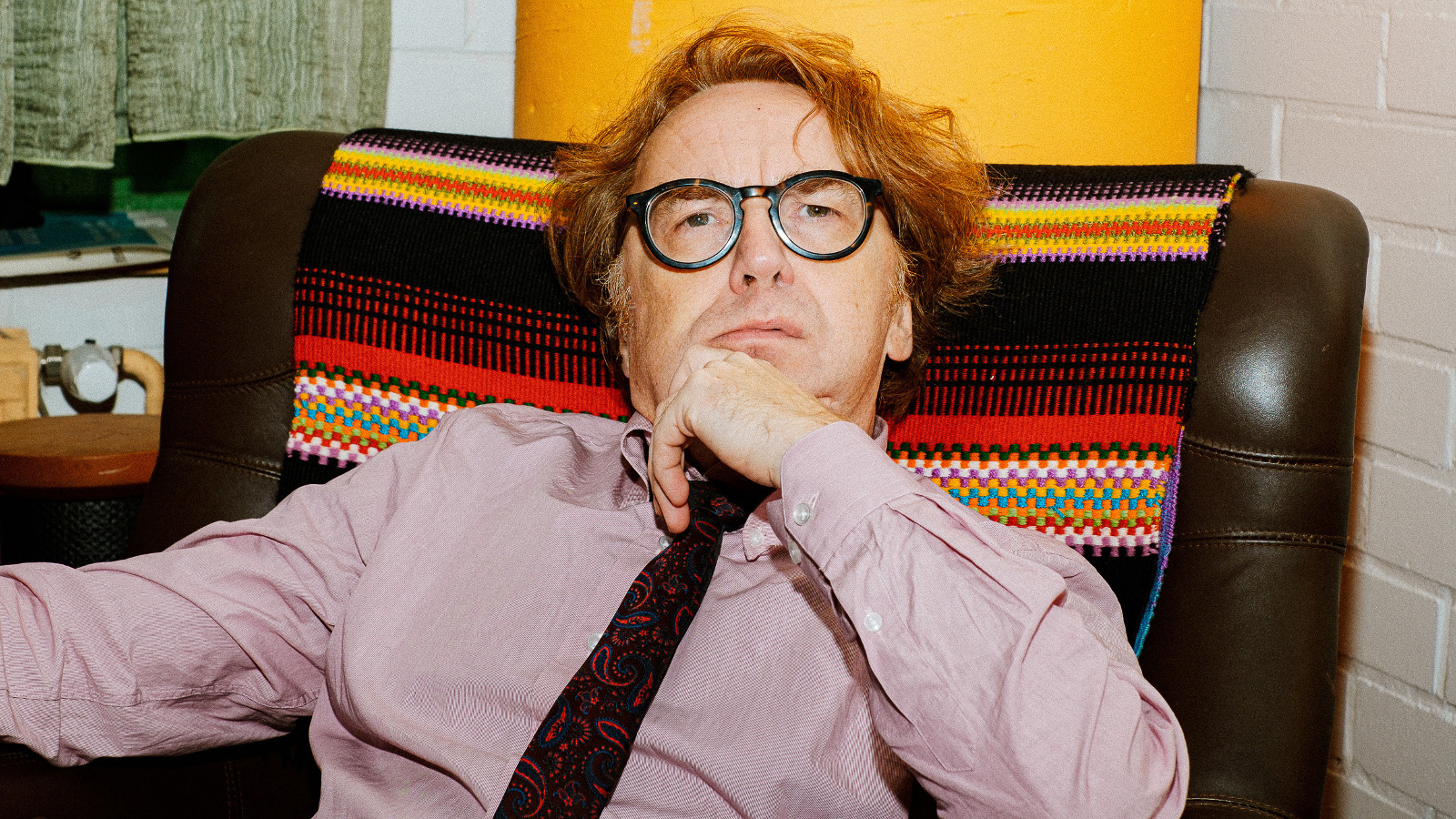 Kuvassa on Pertti Jarla istumassa nojatuolissa puolivartalokuvassa. Hänellä on lilahtava paita ja tumma kravatti. Selkänojassa on tumman ja punaisen raidallinen matto. 