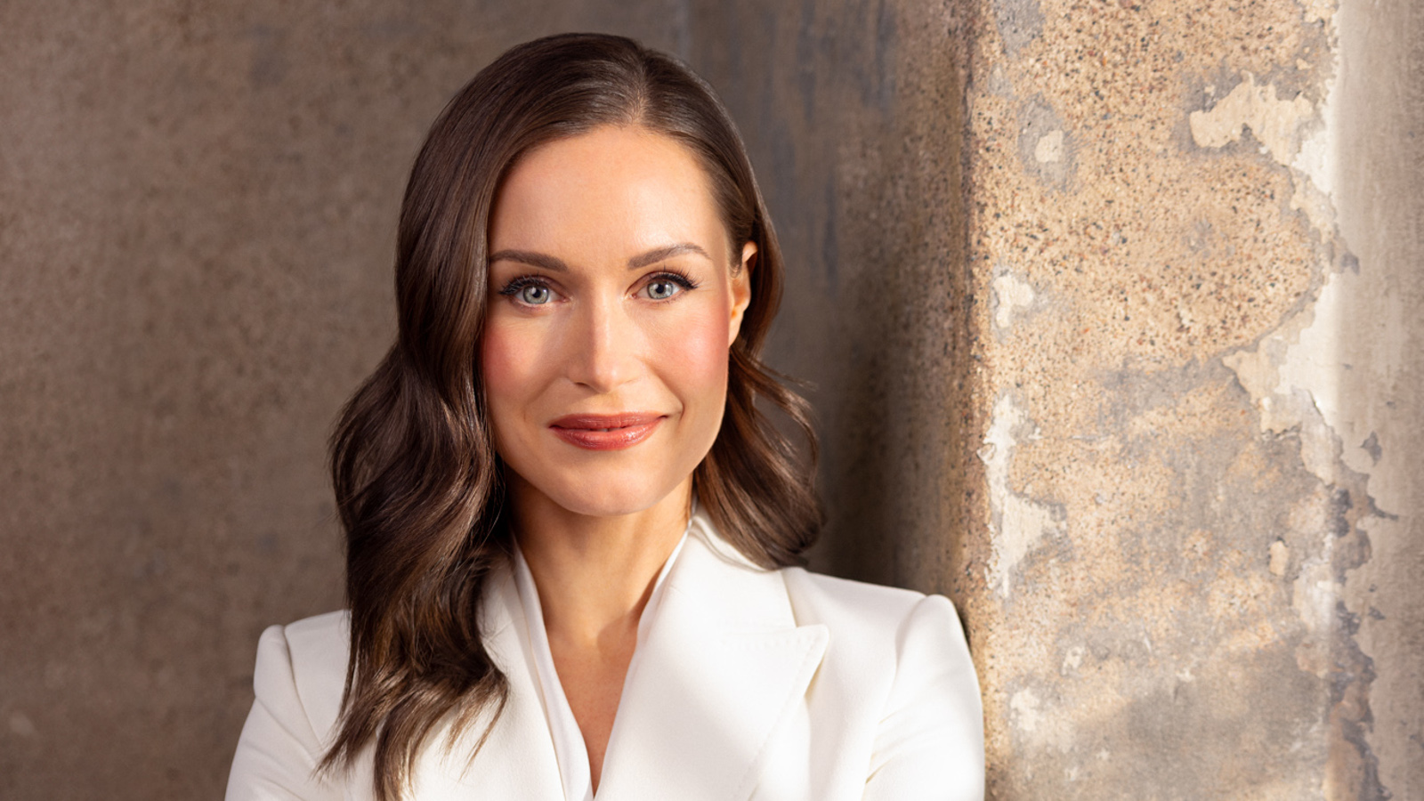 Kuvassa on Sanna Marin puolivartalokuvassa ja hänellä on valkoinen jakku päällään, jossa on v-aukko. Hänellä on pitkät tummat hiukset. Tausta on vaalean ruskeaa kiviseinää.