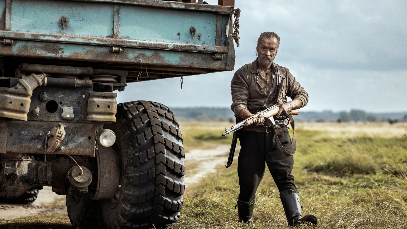 Kuvassa on vasemmalla kuorma-auton korkea takaosa.  Sen vieressä seisoo Jorma Tommila ase kädessään vihreässä maastopuvussa. Taustalla on vihreää ruohoa ja hiekkatietä. 