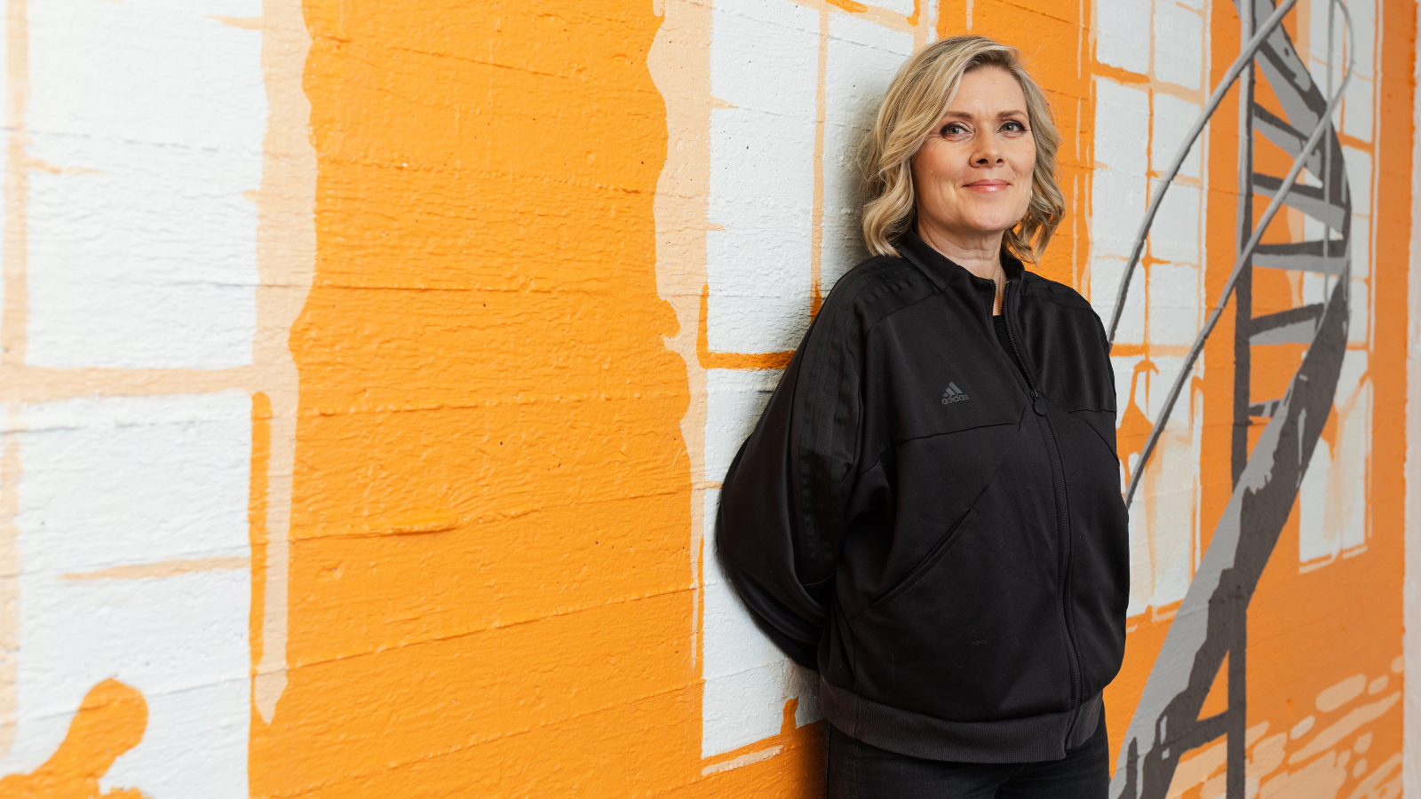 Kuvassa on seinällä on valkoisia kuutioita ja keltaisia leveitä raitoja. Oikealla seinään nojaa taiteilija Ruusa mustassa mekossa. Hänen vieressään oikealla on mustat kierreportaat.   