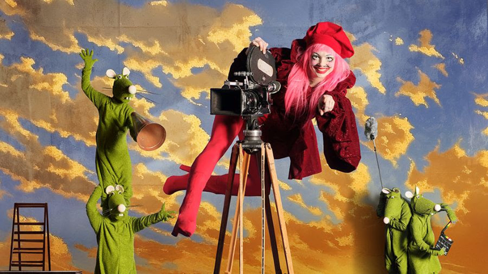 Kuvassa taustalla kelta-harmaita pilviä sinisellä taivaalla. Vasemmalla on kaksi vihreätä hiireksi pukeutunutta hahmo ja toinen seisoo okapäillä. Keskellä on vanhanaikainen filmikamera ja siinä päällä roikkuu nainen punaisissa saappaissa legginsseissä sekä viininpunaisessa takissa. Takana on pieninä kaksi vihreätä hiirtä.