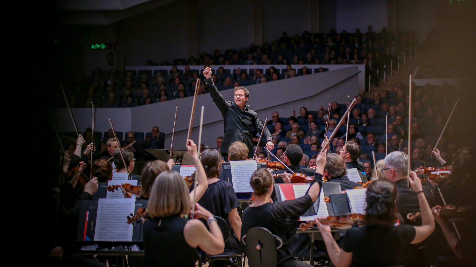 Kuvassa on keskellä Matther Halls johtamassa Tampereen Filharmoniaa.  Orkesterin soittajat ovat selin. Taustalla on osittainn tummaa.