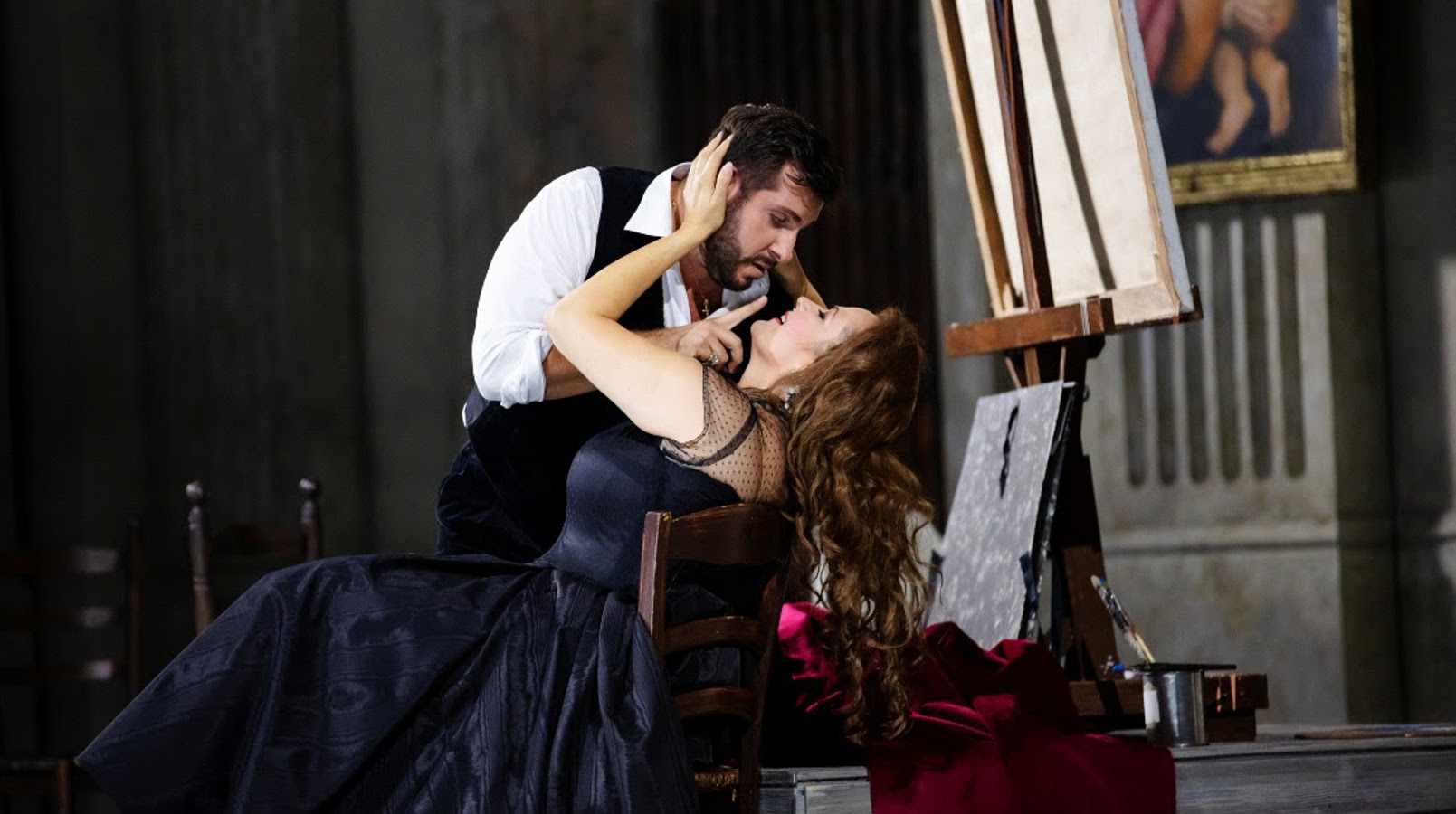 Kuvassa on Toscan roolin laulaja tumman sinisessä pitkässä puvussa istumassa tuolissa ja nojaa selkänojaan.  Hänen ylläpuolellaan on kuvartuneena kasvoja kohti mies valkoisessa puserossa ja mustassa liivissä. Tausta on tumma.