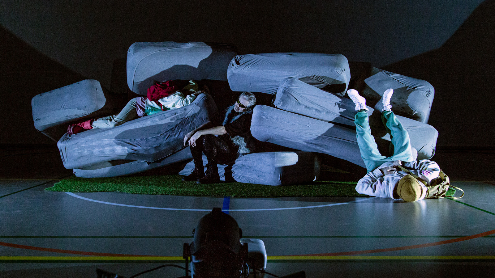 Kuvassa on isojen harmaiden tyynyjen kasa ja välissä on kaksi näyttelijää. Yksi on jalat ylöspäin makaamassa lattilalla oikeassa kulmassa.