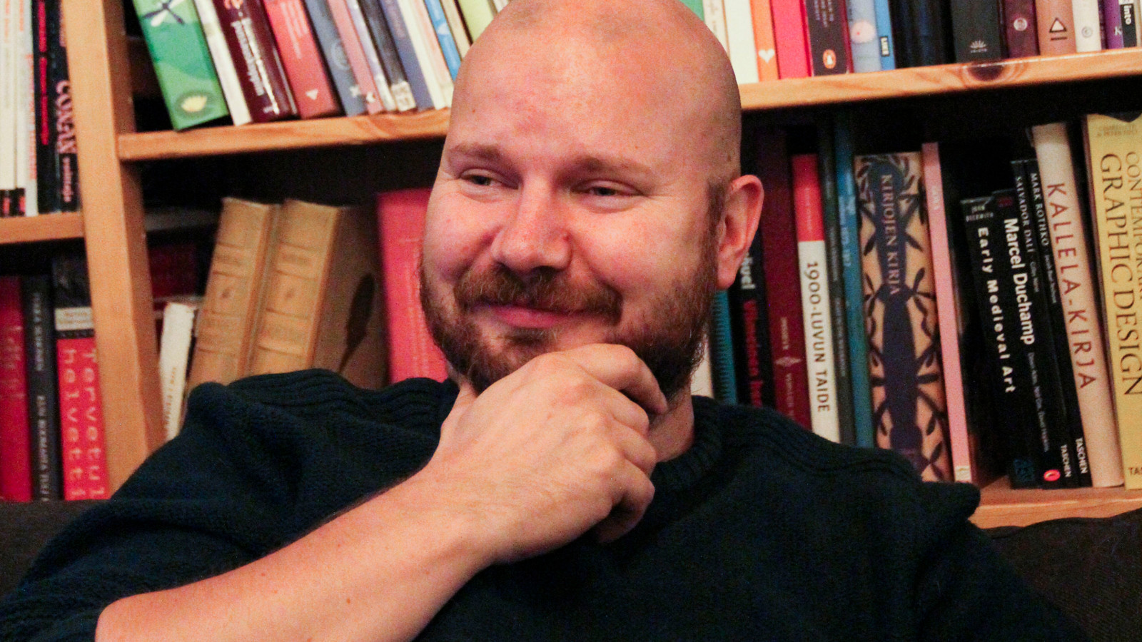 Kuvassa on Tuomo Rämö puolivartalokuvassa istumassa takanaa kirjahyllyä. Hänellä on musta paita ja hän pitää oikeaa kättä leuassaan.