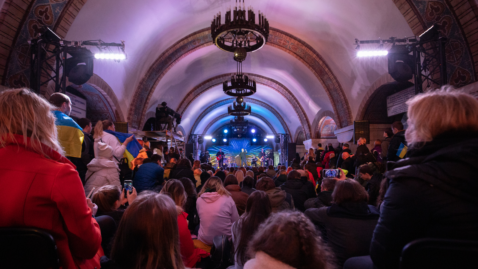 Kuvassa on kaarevaa koristelampullista metrotunnelia ja siellä takana bändi soittamassa. Ihmisiä on selin värikkäissä vaatteissa katsomassa konserttia.    