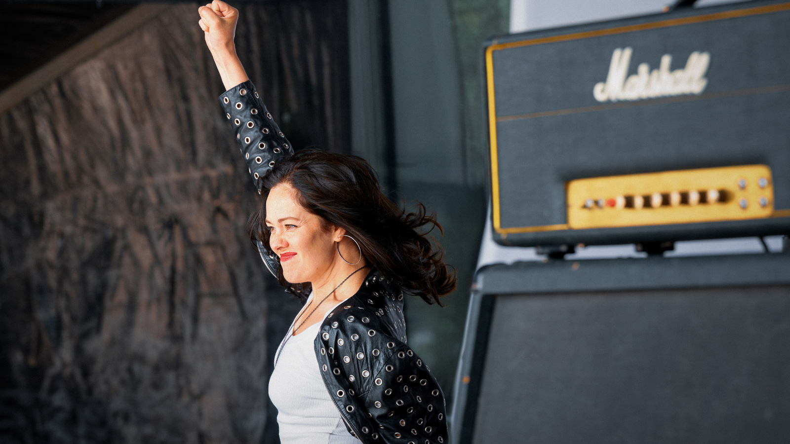 Kuvassa on Oona Airola, joka on lavalla oikea käsi pystyssä valkoisessa puserossa sekä mustassa jakussa. Taustalla näkyy tummaa seinää oikealla on kaiuttimen yläosassa tekstiä ja alemana mustaa kaiutinta.