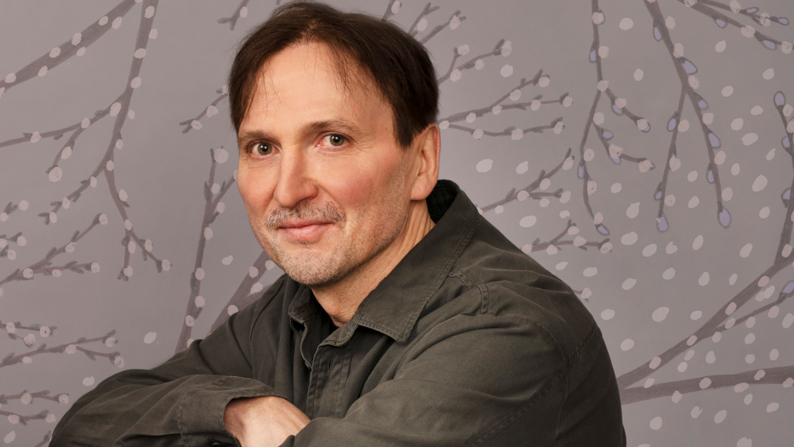 Kuvassa on Tomi Kontio puolivartalokuvassa istumassa ruskeassa paidassa. Taustalla on harmaalla pohjalla pajunkissatapettia.