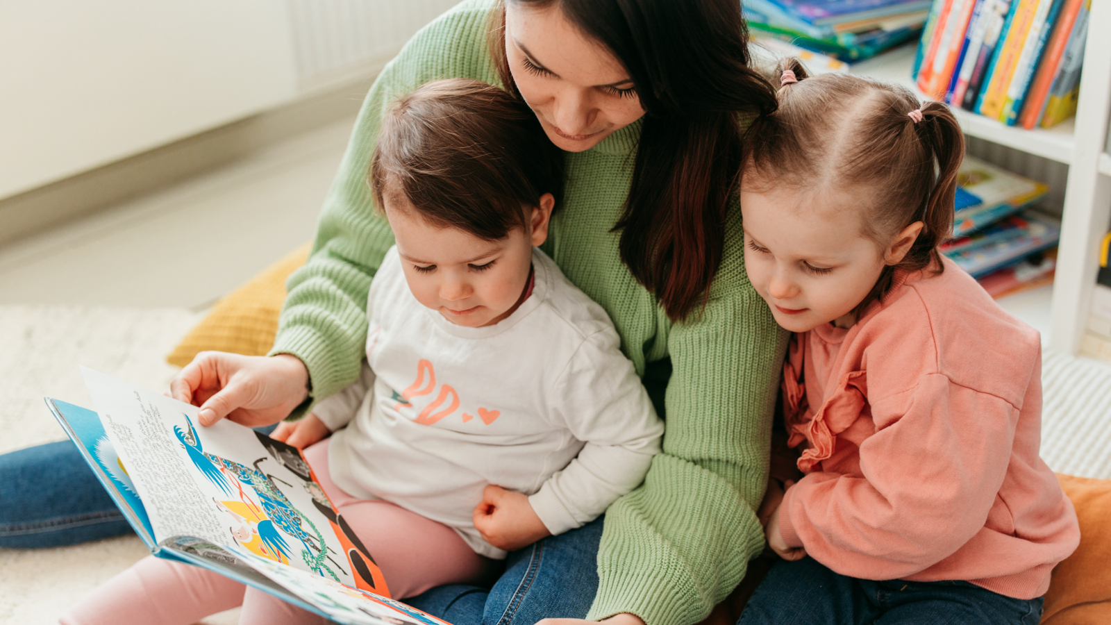 Kuvassa on nuori nainen, joka lukee kirjaa kahdelle sylissään olevalle lapselle. He ovat noin neljä- ja kuusivuotiaita. 