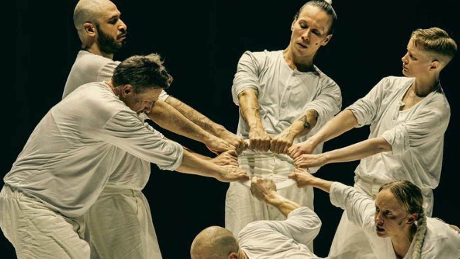 Kuvassa on viisi miestanssijaa ja yksi näinen vaaleissa kangasasuissa ja he pitävät kahdella kädellä kiinni vaaleasta renkaasta. Edessä oleva tanssija on alhaalla kumartuneena.