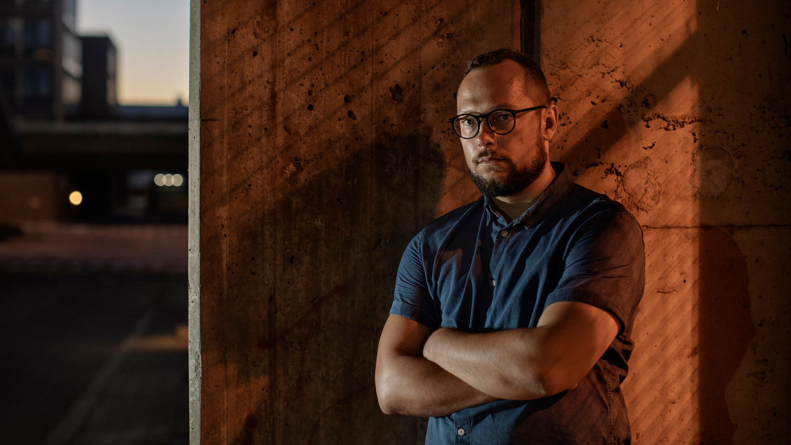  Kuvassa on Paavo Teittinen seisomassa kädet ristittynä rinnalle.  Hänellä on sininen pikeepaita päällään. Taustalla on ruskea seinä ja vasemmalla näkyy hämärää kerrostaloaluetta.