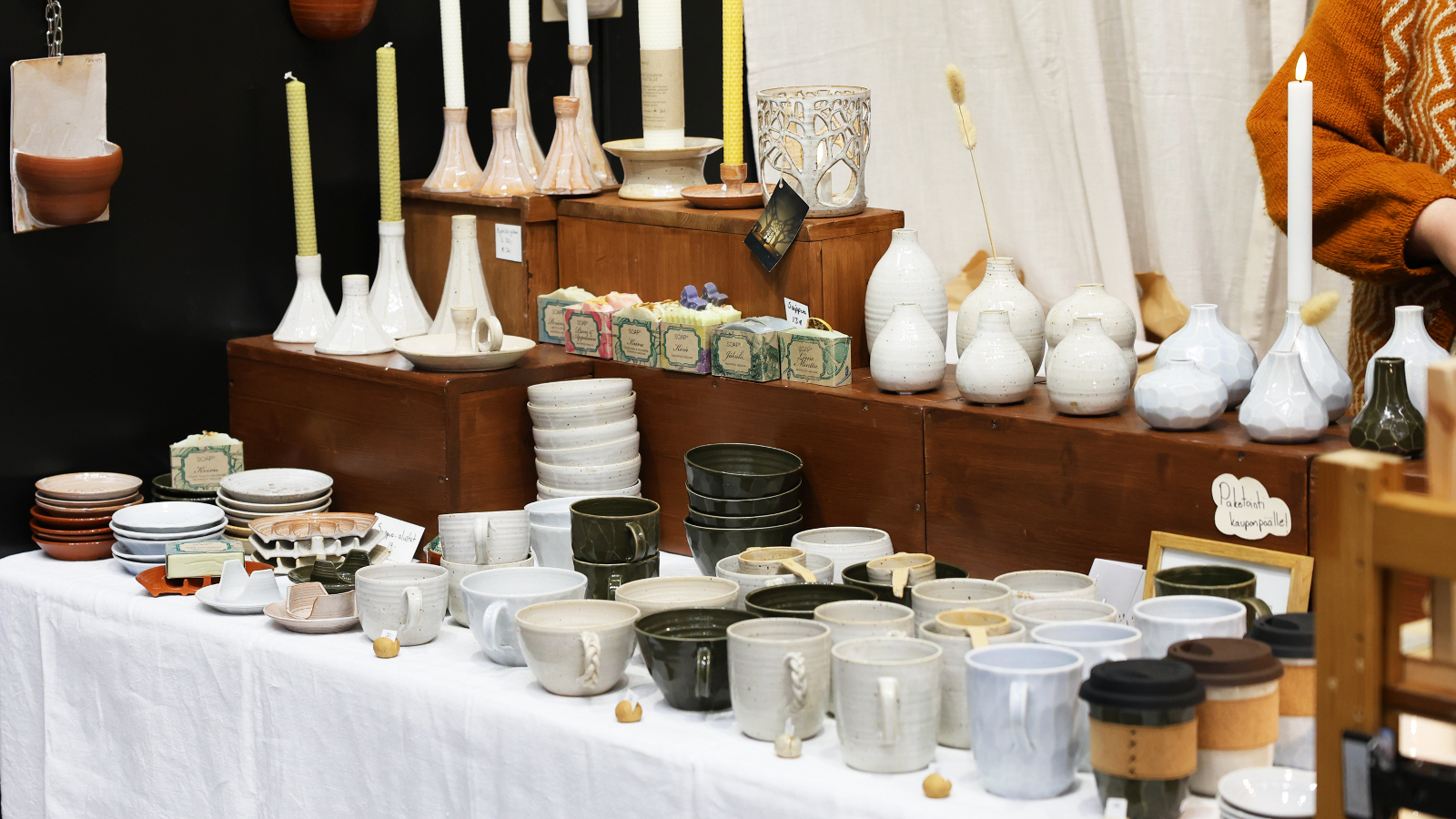 Kuvassa on pöydällä keraamisia kuppeja ja ruskealla hyllyllä korkeita kynttilöitä ja matalia kynttilöitä.