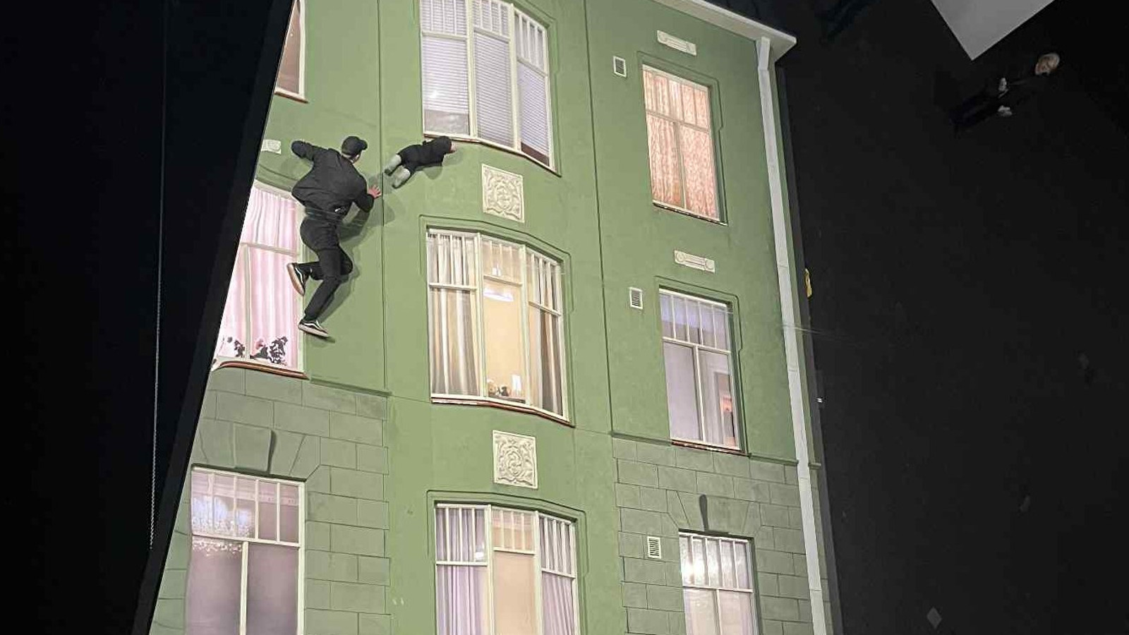 Kuvassa on molemmin puolin mustaa ja keskellä on vaalean vihreä vanha kerrostalo, jossa on kolmeen osaan jakautuneet ikkunoita kolmessa rivissä. Vasemmalla ylhäällä on nuori mies mustassa puvussa mukanaan vauva kiipeämässä seinällä.