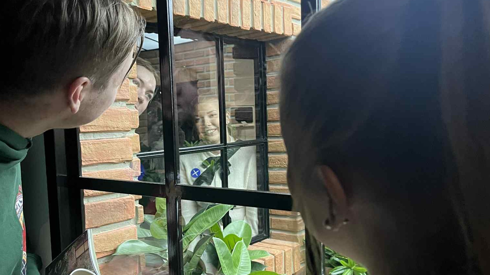 Kuvassa on vasemmalla nuori mies kurkistamassa ikkunasta ja oikealla takaa nainen. Ikkunasta näkyy kulmittain keltaista tiiliseinää ja ruudullinen ikkuna. Sieltä näkyy katsojien kuvat.