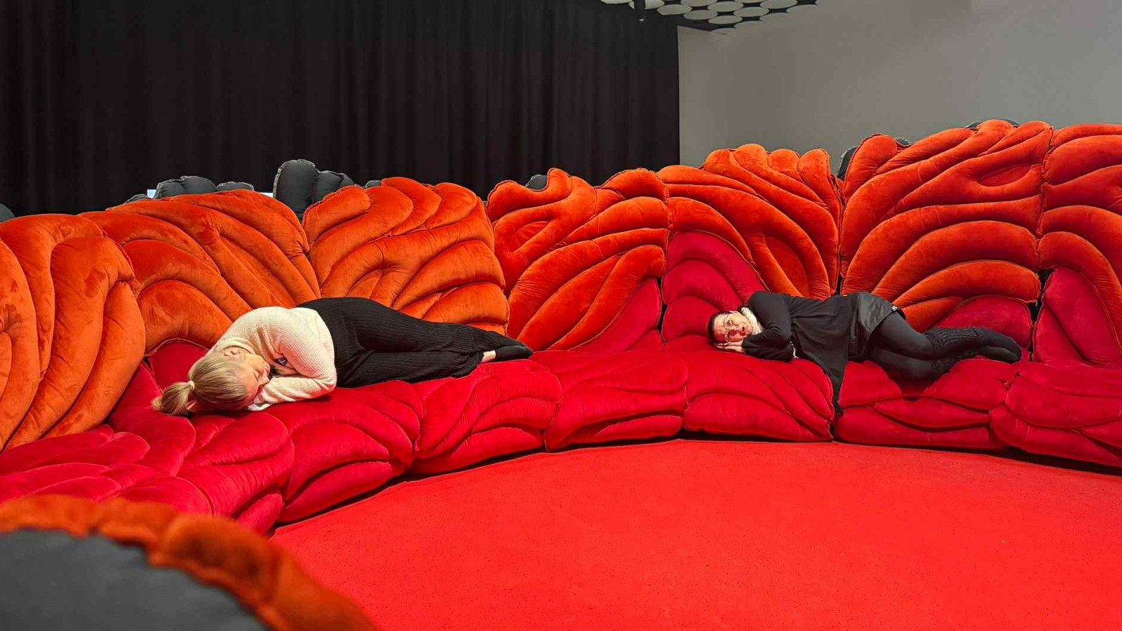 Kuvassa on iso kaareva, punainen sohva ja keskellä punaista lattiaa. Tausta on tumma. Sovalla makaa kaksi naista.