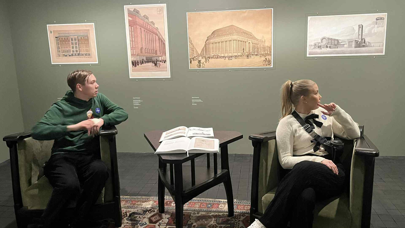 Kuvassa on vihertävällä seinällä neljä piirroskuvaa, joissa on Helsingin keskustan rakennuksia. Vasemmalla on tummassa on kaarevassa, puisessa nojatuolissa istuu vinottain katsomassa teoksia vihreässä hupparissa nuori mies. Oikealla on istumassa ja kääntyneenä katsomaan teoksia nuori nainen valkoisessa puserossa ja mustissa pitkissä housuissa.