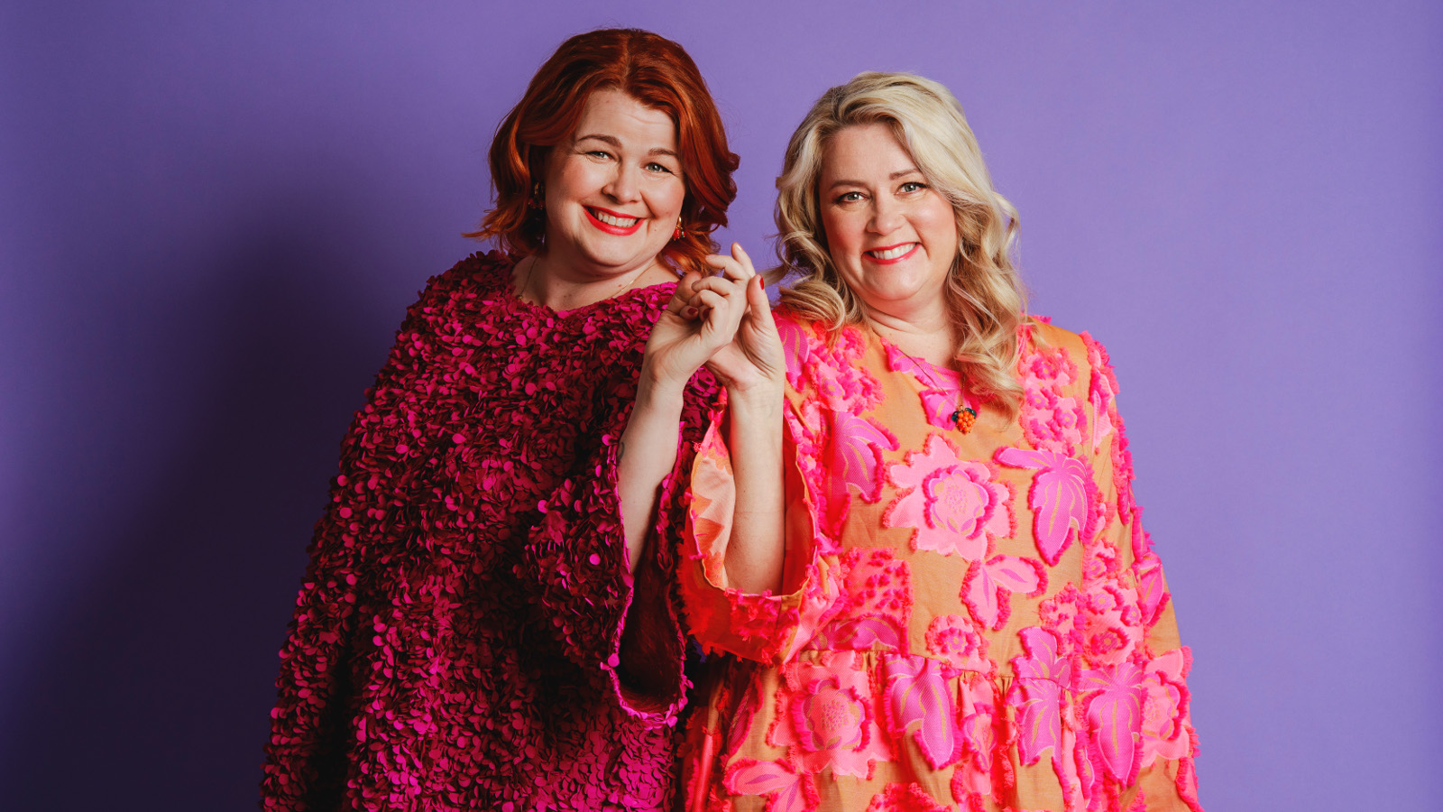 Kuvassa on vasemmalla seisomassa Ani Kellomäki viininpunaisessa mekossa  ja Tiia Koivusalo pinkissä mekossa. Tausta on lila.