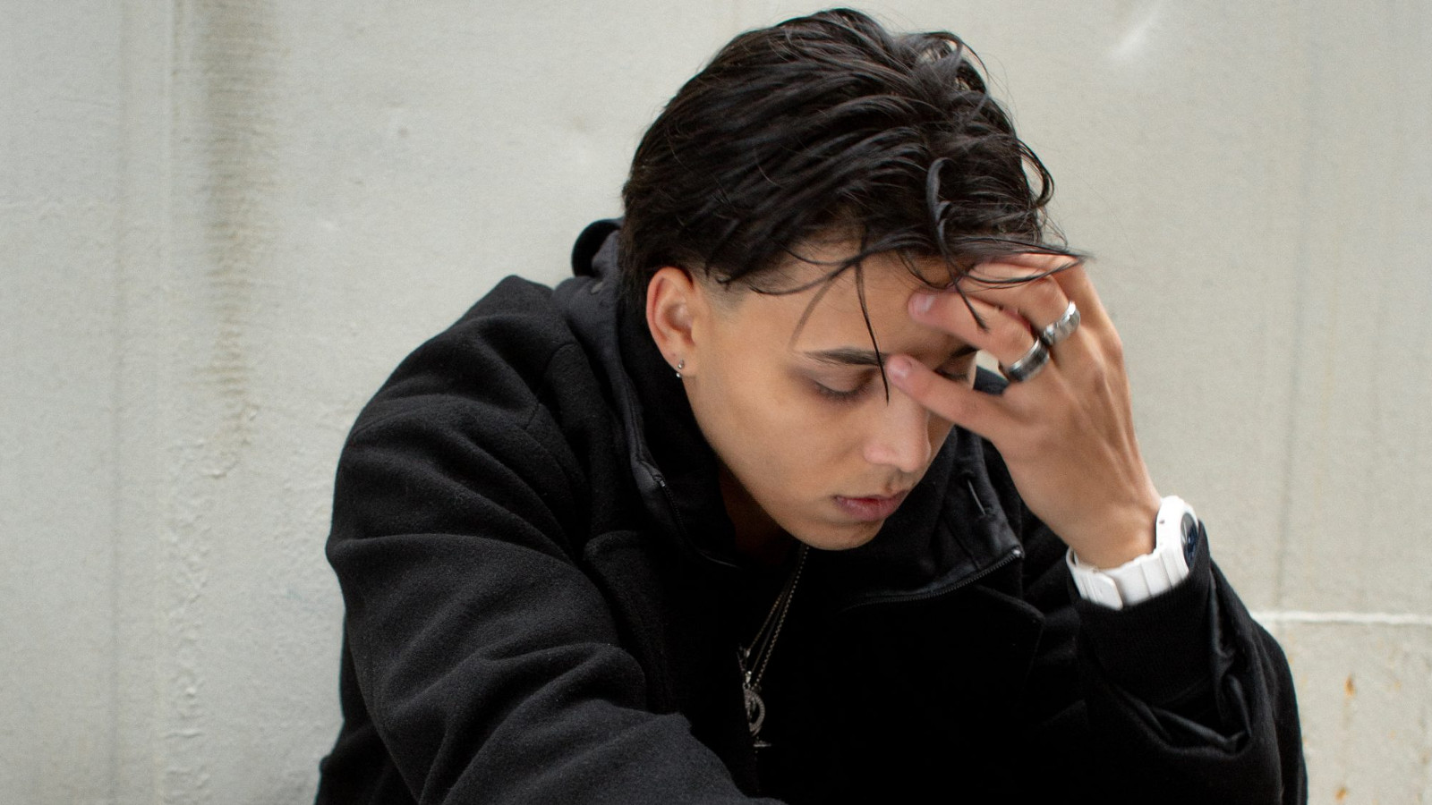 Kuvassa on Averagekidluke istumassa puolivartalokuvassa kuvartuneena. Hän pitää vasemmalla kädellään otsaansa. Tausta on vaalean harmaa.