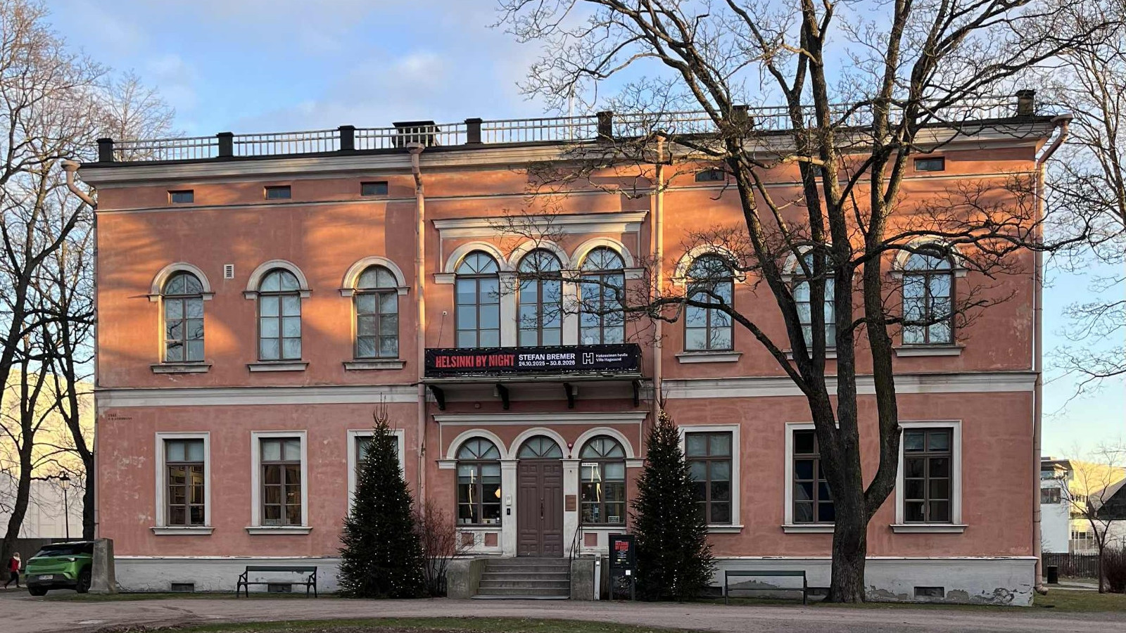 Kuvassa on Hakasalmen huvila, joka on kaksi kerroksinen ja väriltään oranssi. Molemmissa kerroksissa on rivi ikkunoita, joissa on yläkaari. Lähellä on lehdettömiä puita.