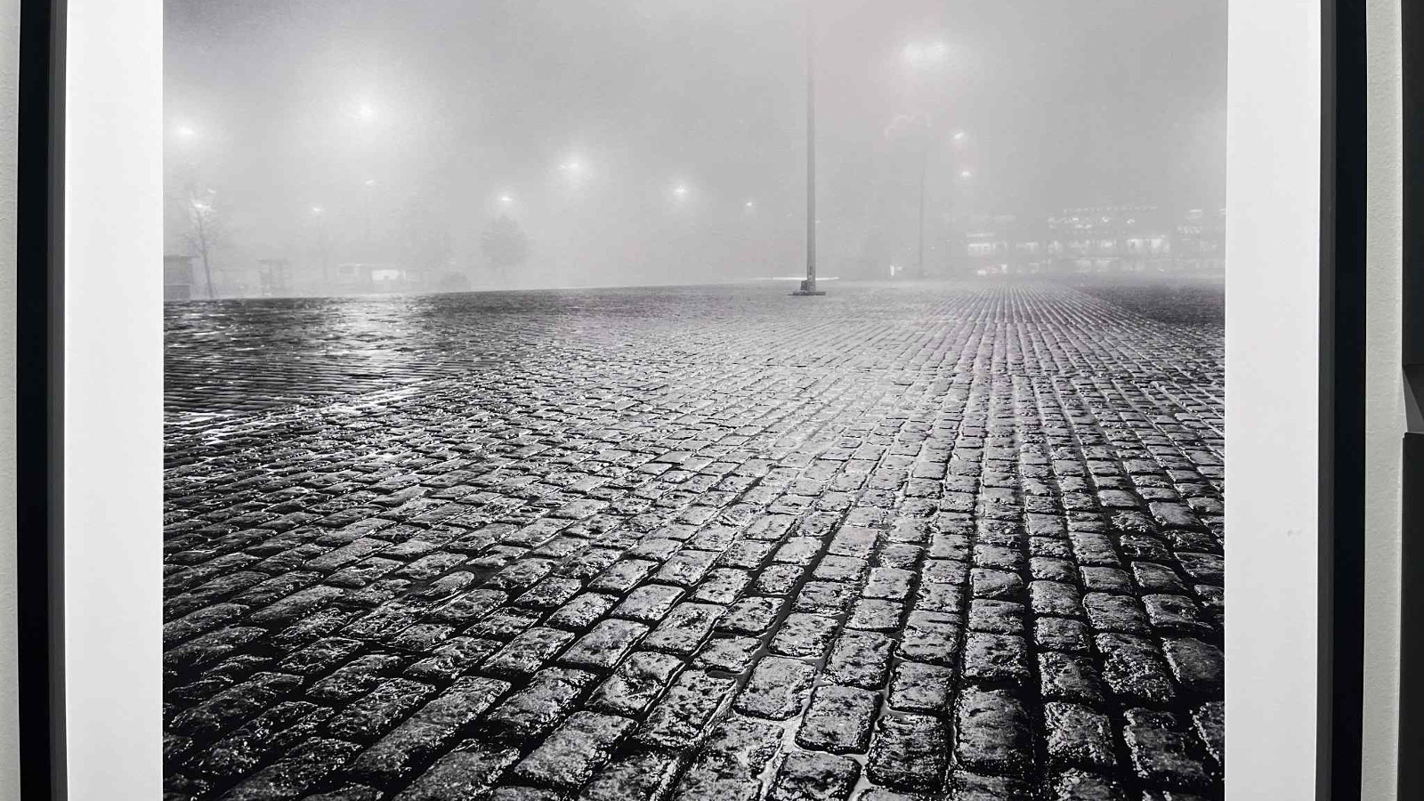 Kuvassa on musta-valkoinen valokuva öisestä torista ja sen märistä katutiilistä. Taustalla on sumua ja himmeitä katuvaloja.
