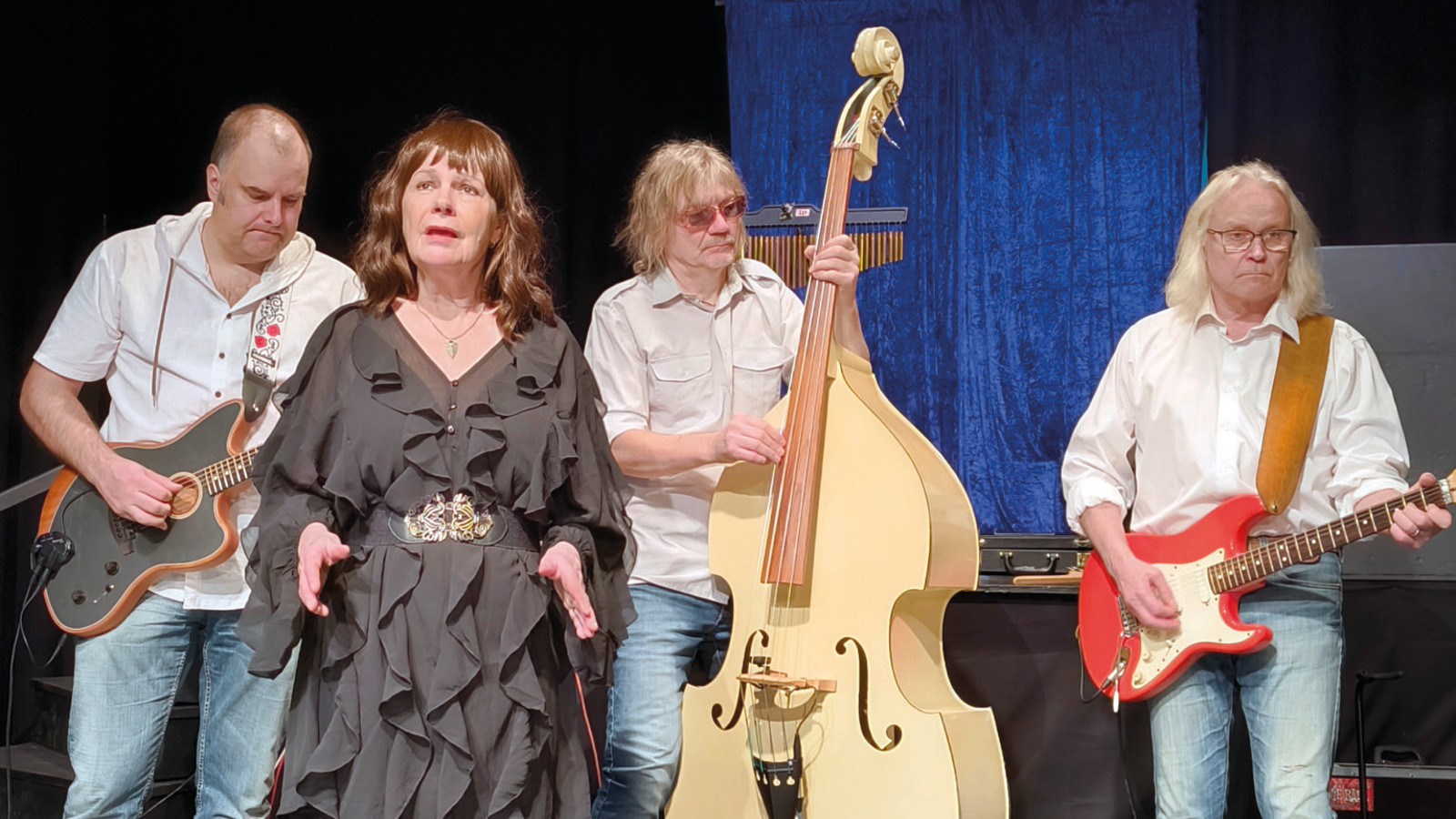 Kuvassa on vasemmalta kitaristi ja hänen vieressään edessään nainen ruskeassa rimpsutetussa mekossa ja keskellä on vaaleaa bassoa soittava mies. Oikealla on kitaraa soittava mies valkoisessa paidassa ja farkuissa.