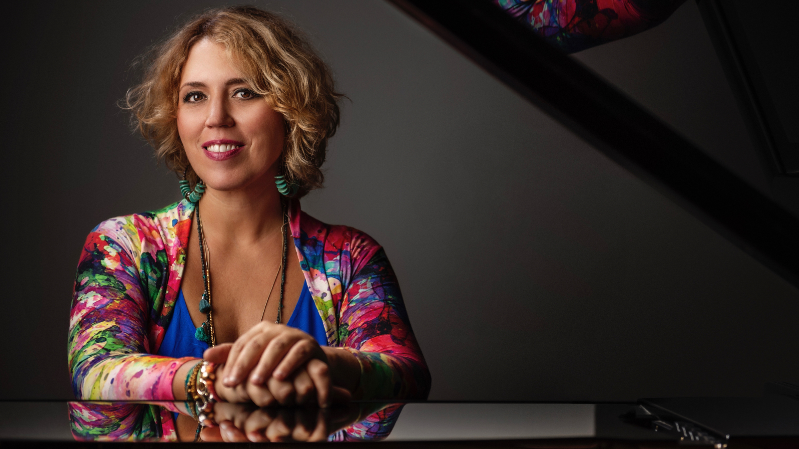 Kuvassa on vasemmalla Gabriela Montero istumassa flyygelin takana ja pitää käsiään kiiltävän flyygelin kannen päällä. Oikealta kaartuu flyygelin kansi osittain pään yli. Hänellä kukallinen puna-sini-valkokirjava pusero. Tausta on tumma.