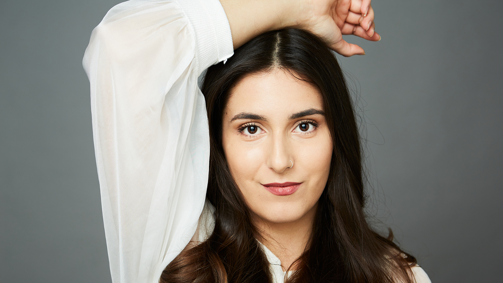 Kuvassa on Iris Candeleria puolivartalokuvassa ja hänellä on pitkt tummat hiukset. Hänellä on valkoinen pitkähihainen pusero ja hän pitää oikeaa kättään päälaellaan. Tausta on harmaa.