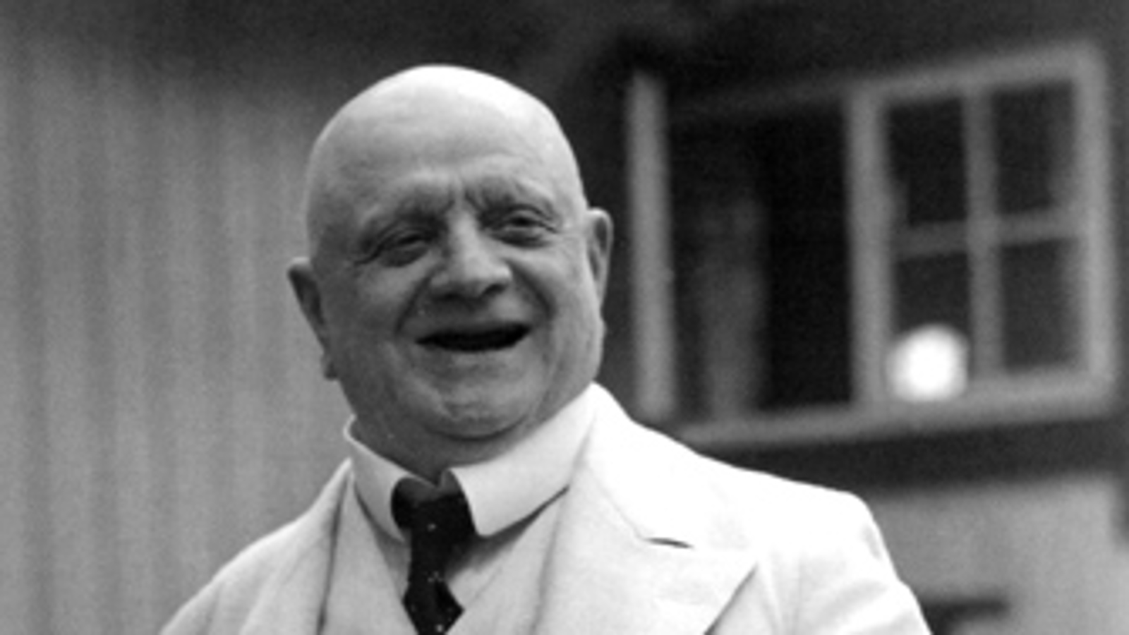 Kuvassa on Jean Sibelius kaljupaisenä puolivartalokuvassa ja hänellä on valkoinen takki ja paita sekä m usta solmio. Taustalla näkyy talon seinää ja ikkunaa. Kuva on musta-valkoinen.