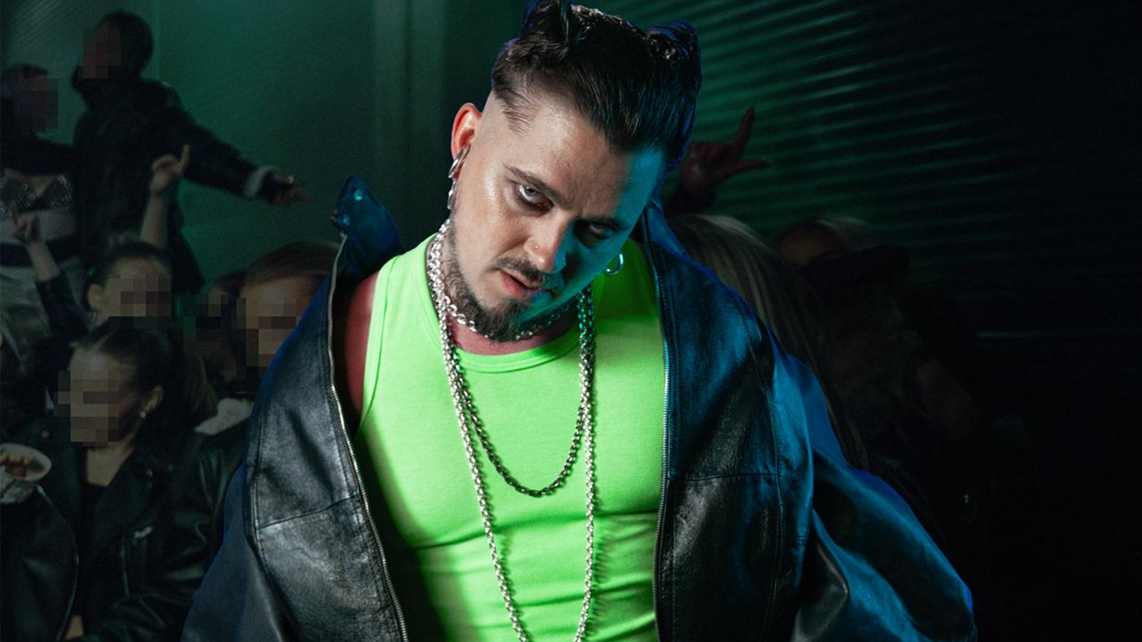 Kuvassa on Käärijä puolivartalokuvassa. Hänellä on hihaton vaalean vihreä t-paita ja musta nahkatakki olkapäillään kevyesti. Taustalla on tummahkoa ja alempana näkyy yleisön kasvoja.