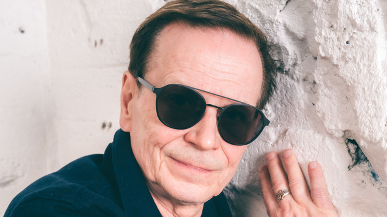 Kuvassa on Kake Randelinin kasvot. Hänellä on aurinkolasit päässään ja hän pitää oikeaa kättään vaaleassa tiilisessä seinässä.