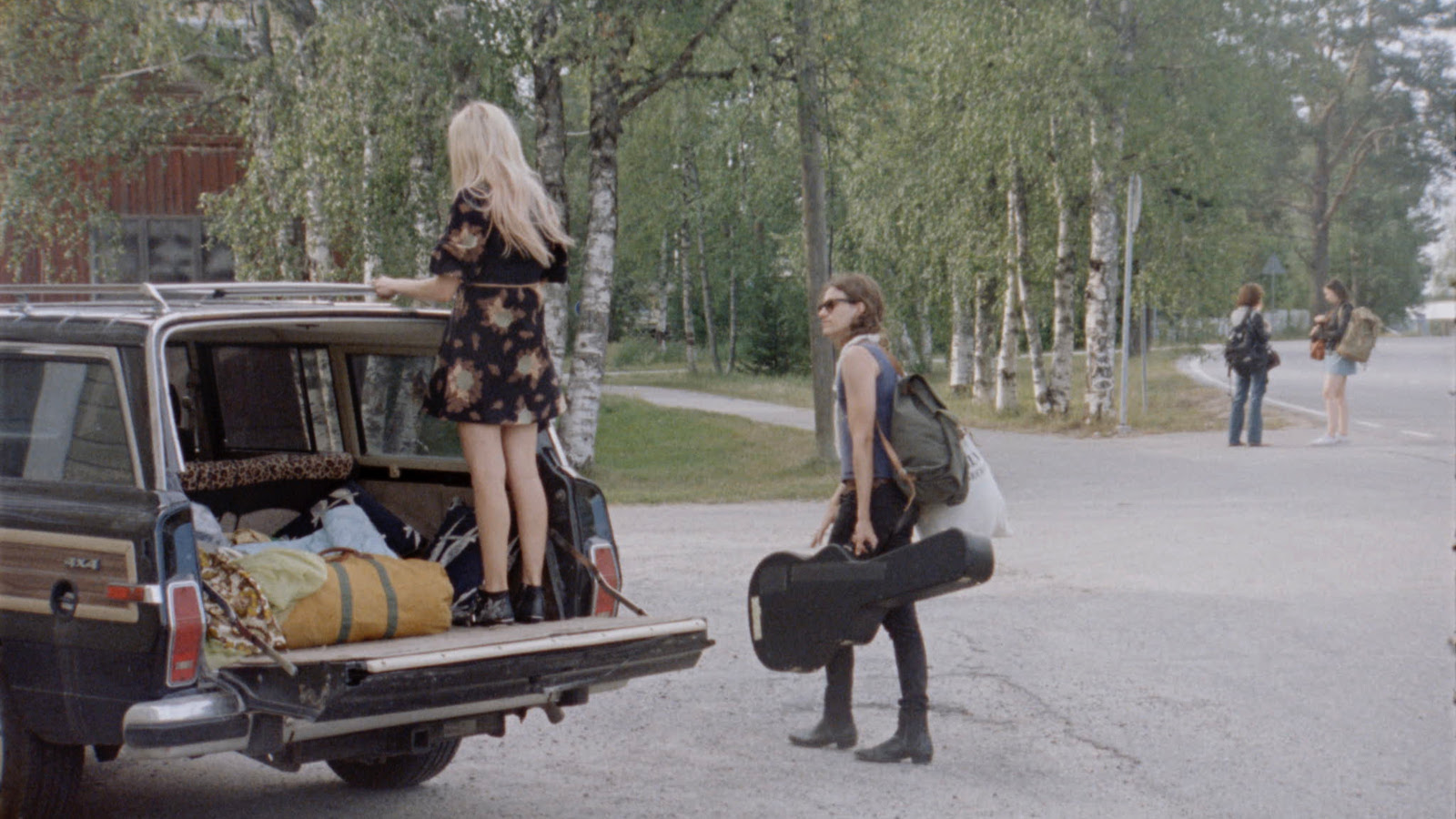 Kuvassa on vasemmalla auton takakontti auki ja sen päällä seisoo reunalla selin Alina Tomnikov. Taustalla on koivumetsää ja asfaltilla oikealla kävelee poispäin nuori mies.