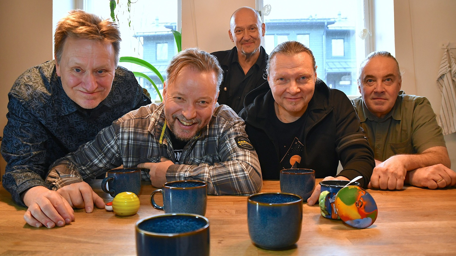 Kuvassa ovat yhtyeen viisi jäsentä vaalean ruskean pöydän ääressä istumassa eri värisissä paidoissa. Edessä on sinisiä mukeja.