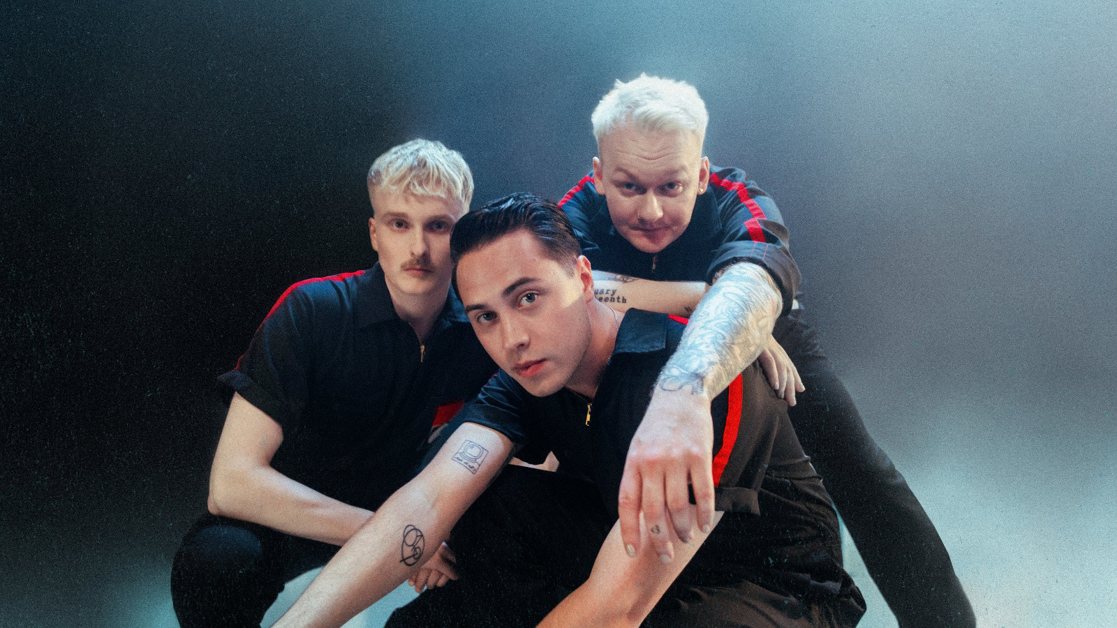 Kuvassa ovat Kuumaa-yhtyeen kolme jäsentä istumassa tiiviisti yhdessä. Heillä on tummat t-paidat päällään. Tausta on tumman sininen.