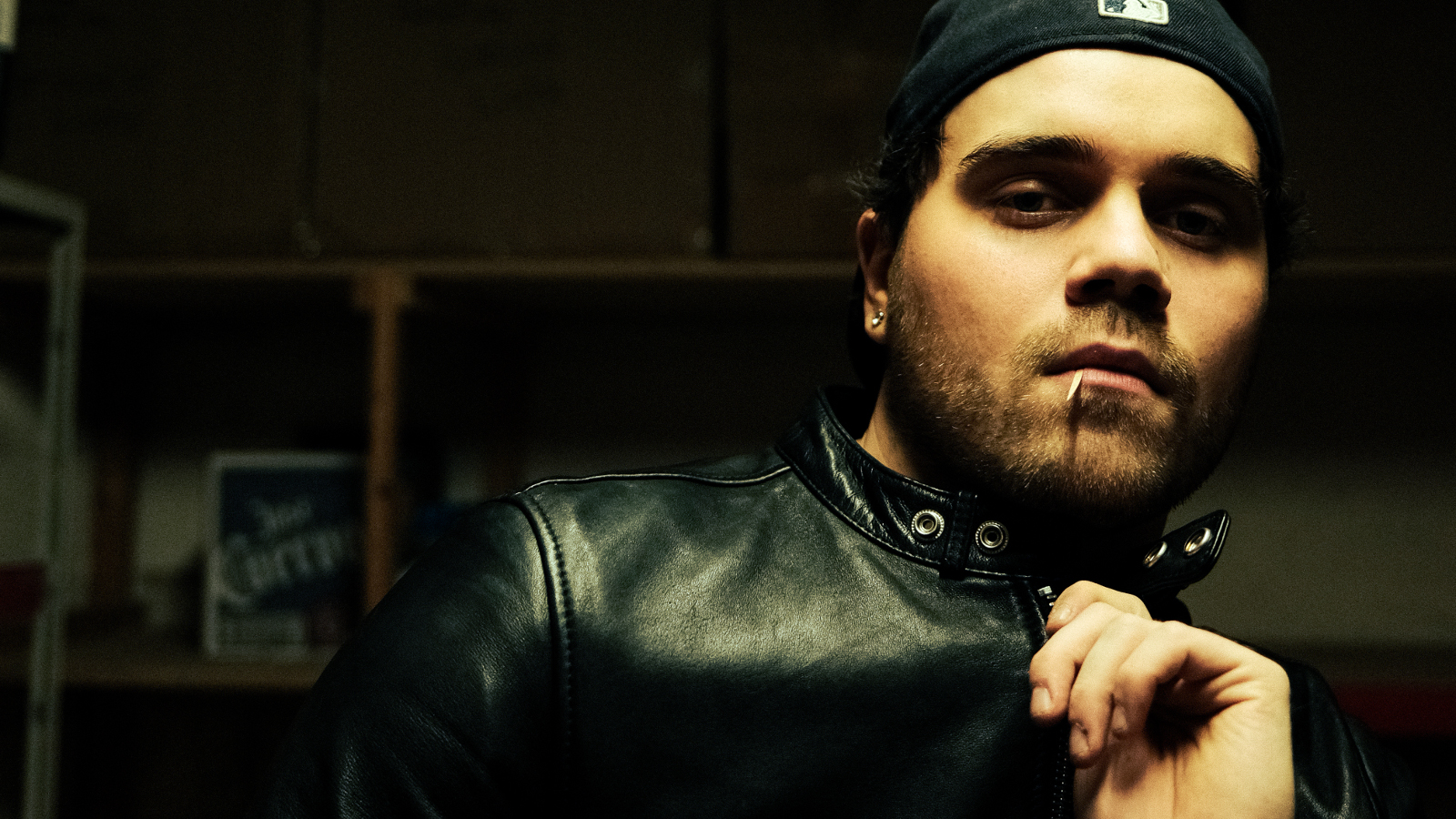 Kuvassa on oikeassa reunassa puolivartalokuvassa ja hänestä näkyy mustalla pohjalla kasvot ja vasenta kämmentä. Hänellä on musta nahkatakki päällään.