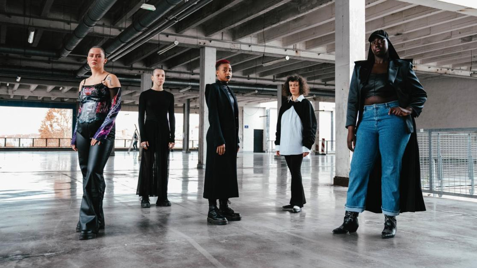 Kuvassa on ryhmässä viisi jäsentä ja he seisovat. Heillä on mustat vaatteet. He ovat matalassa tilassa, jossa on putkistokattoa ja kiiltävää, harmaata lattiaa. Taustalla on isot ikkunat. 