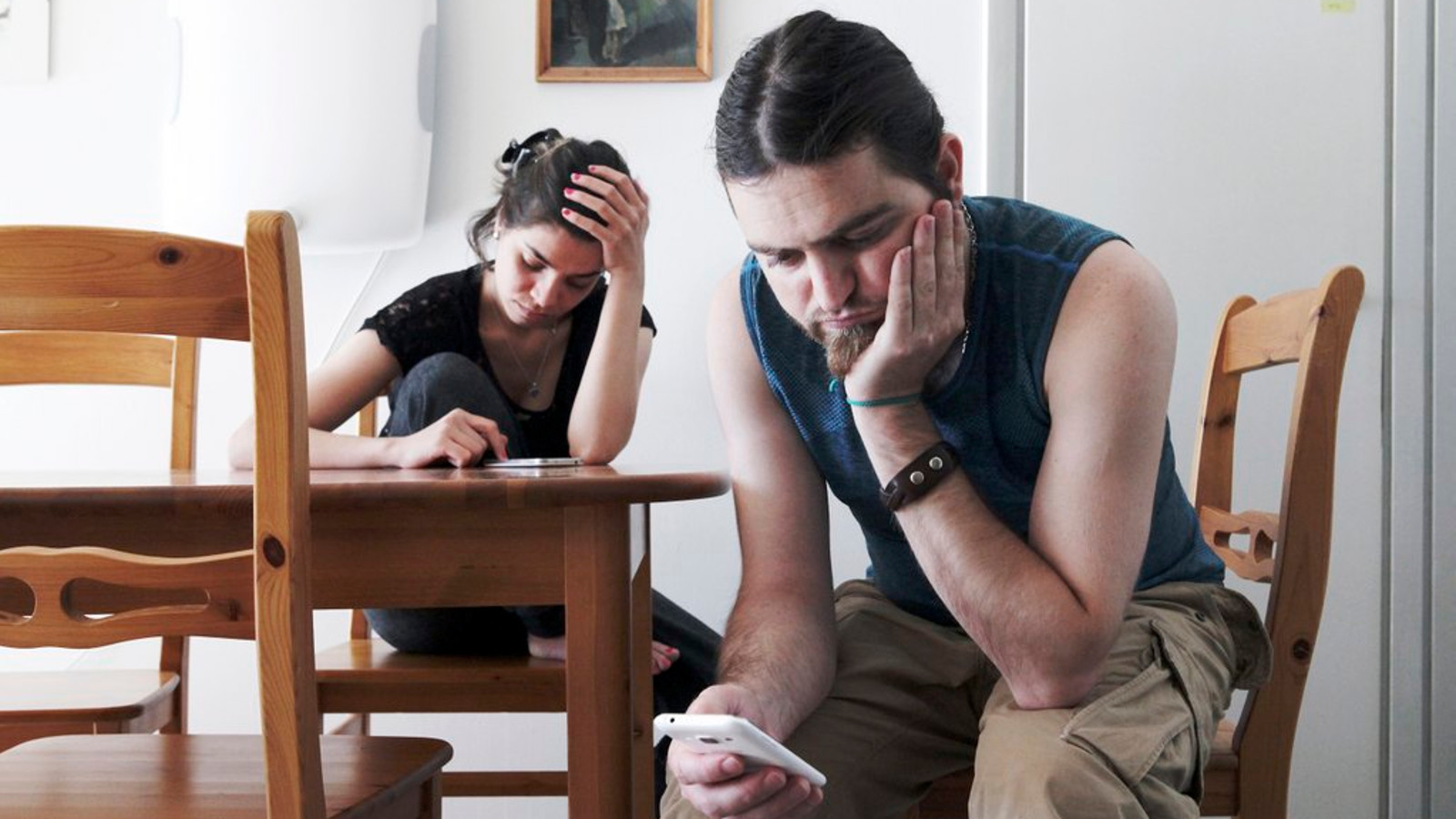 Kuvassa on etualalla oikealla nuori mies istumassa, nojaa vasemmalla kädellään poskeensa ja katsoo oikeassa kädessään olevaa kännykkää. Pöydän takana istuu nuori nainen ja katsoo myös kännykkäänsä. 