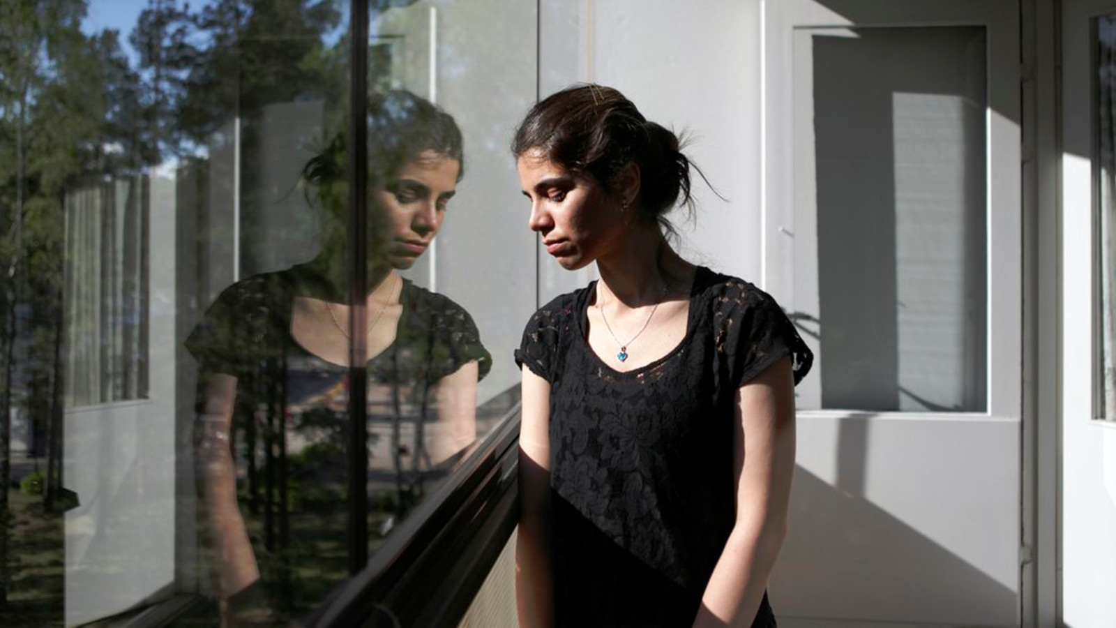 Kuvassa on nuori nainen seisomassa lasitetulla parvekkeella, josta heijastuu hänen oma kuvansa. Hän katsoo alaspäin. Ikkunasta heijastuu vaaleaa taloa ja mäntyjä. Hänellä on musta t-paita.