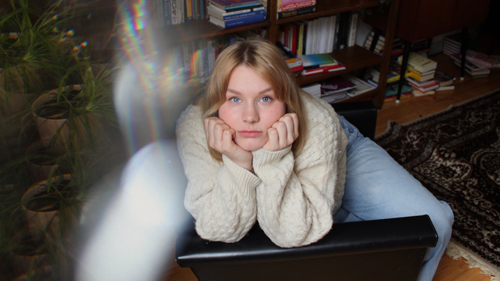 Kuvassa on Riina Tanskanen makaavassa asennossa pöydän päällä valkoisessa villapuserossa. Taustalla on tumma kirjahylly.