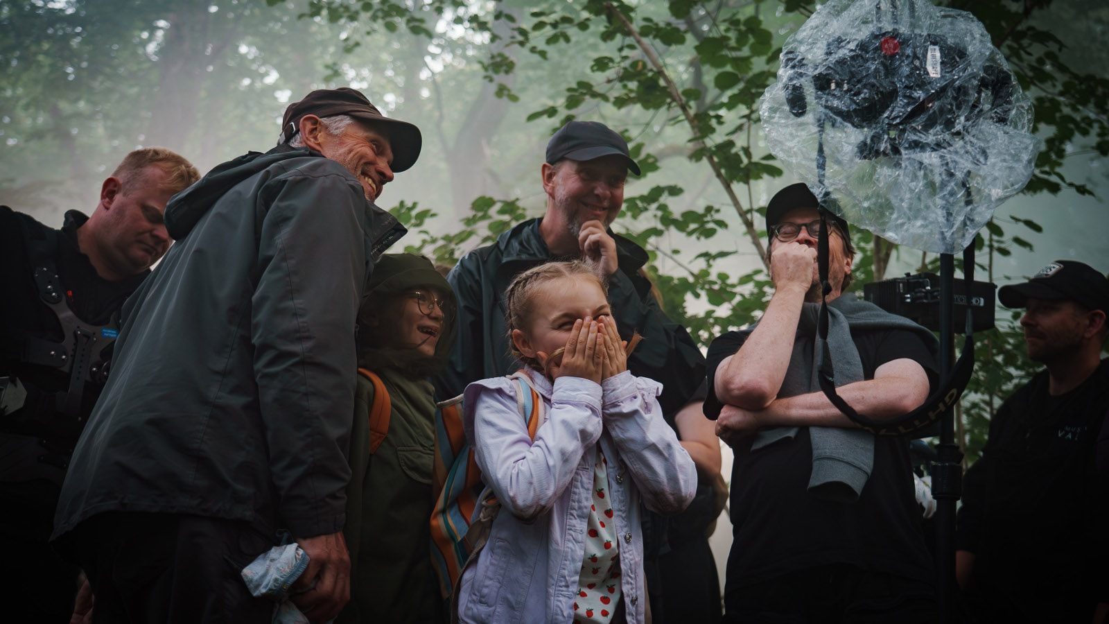 Kuvassa on keskellä vaalean sinisessä farkkutakissa Siiri Virtanen ja hänen ympärillään on kuvausryhmän kuusi henkilöä tummissa ulkovaatteissa. Yläpuolella on vihreitä lehtipuun oksia ja kaikki seisovat puun alla.