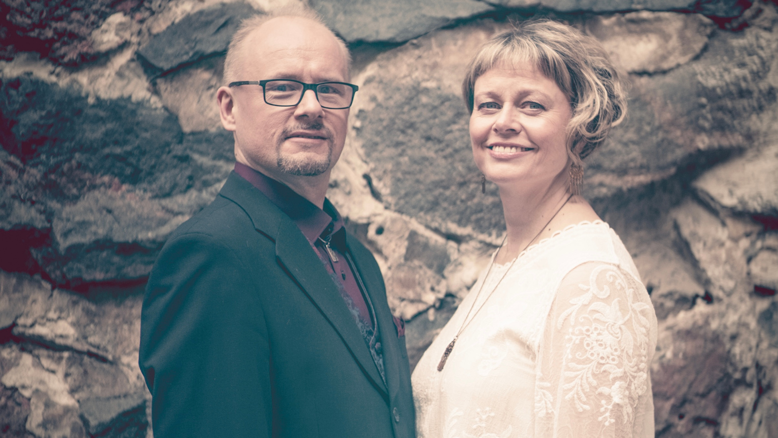 Kuvassa on vasemmalla Tapio Lappalainen mustassa takissa ja Sanna Lappalainen vaaleassa puserossa. Taustalla on kiviseinää, jossa on suuria, tummia kiviä ja vaaleita välimuurauksia.