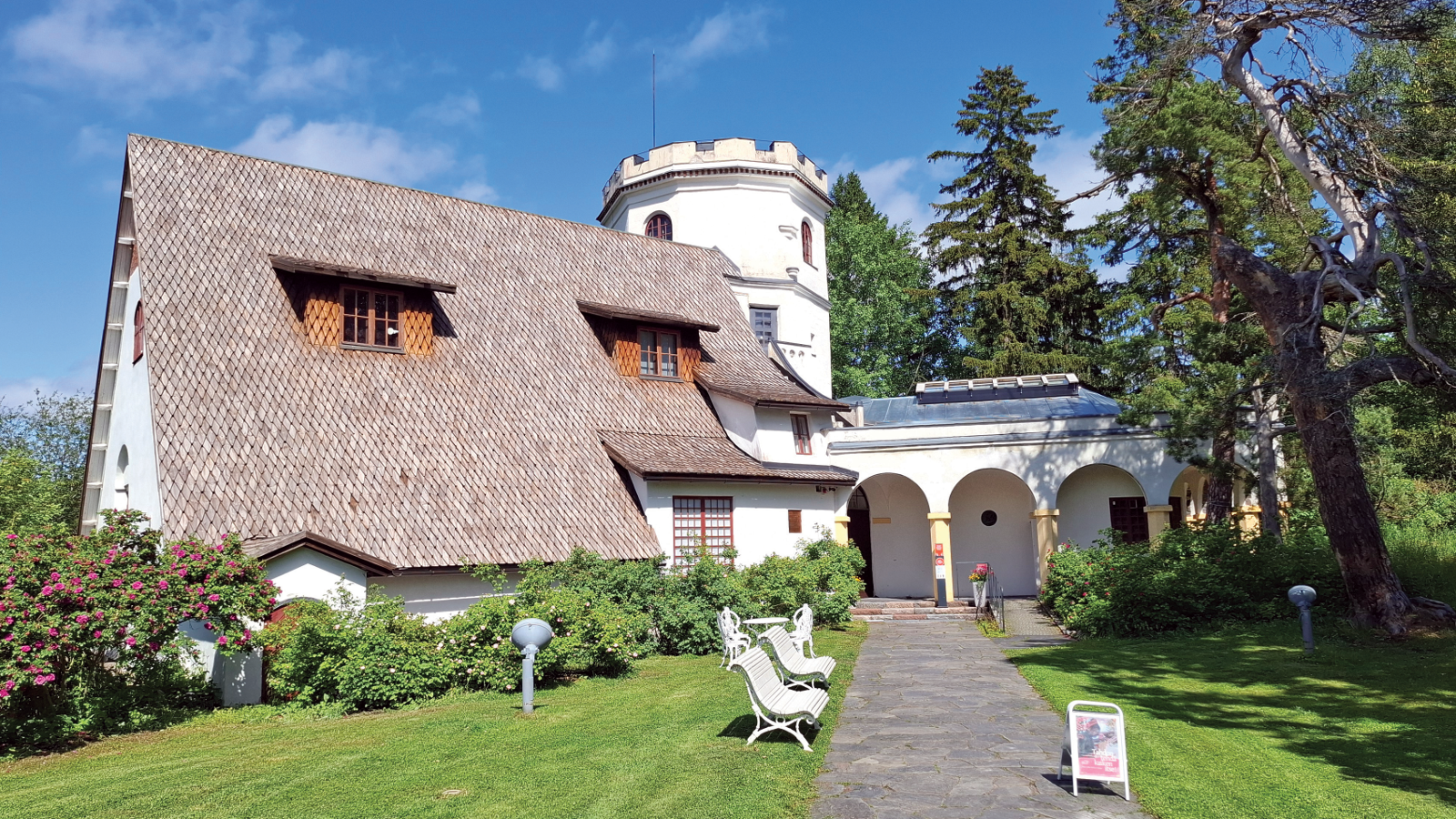 Kuvassa on Tarvaspään päärakennus, jossa on terävä ja alas asti ulottuva harmaa katto, jossa on kaksi kattoikkunaa. Takana valkoinen torni sekä matala lisärakennus kaaripylväikkö. Edustalla on nurmikkoa ja oikealla puita.