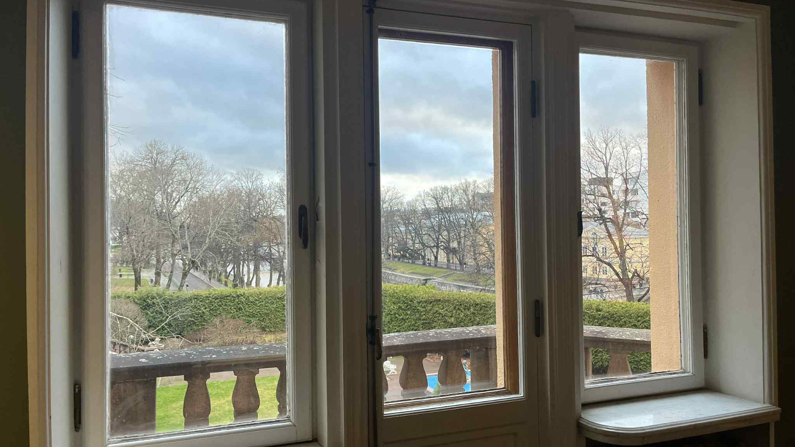 Kuvassa on näkymä kolmiosaisesta ikkunasta, josta näkyy maisemaa, jossa on vihreää pensasaitaa ja lehdettömiä puita sekä kivistä parvekkeen kaidetta.