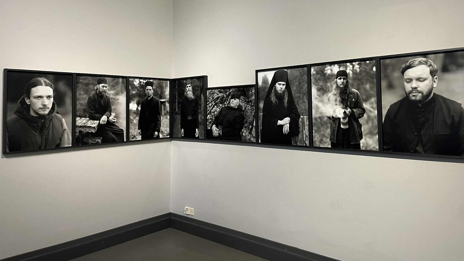 Kuvassa on keskellä seinää valokuvia, jossa on musta-valkokuvia munkeista mustissa vaatteissa. Kuvat jatkuvat kulman yli samalla korkeudella.