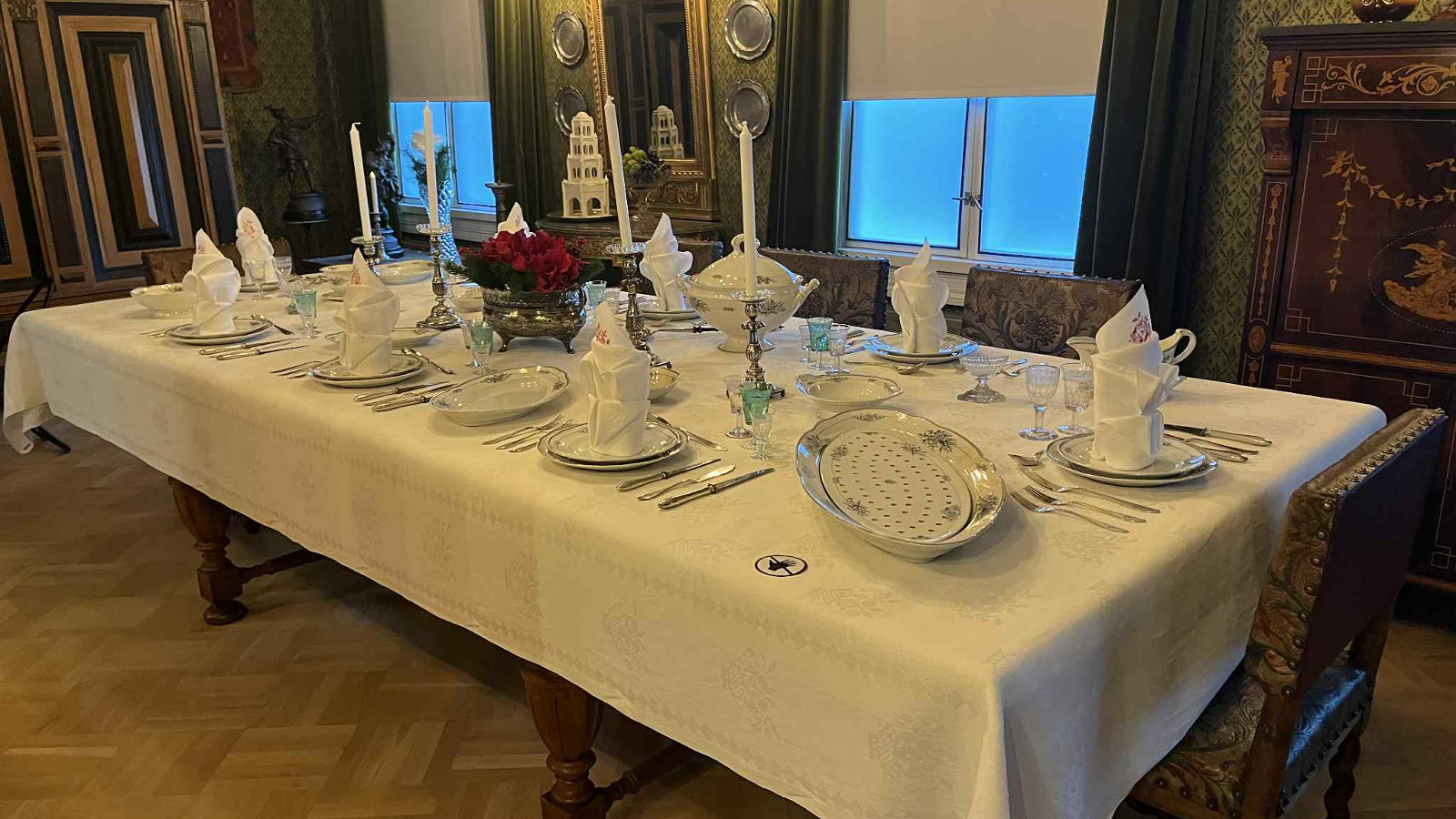 Kuvassa on valkoisella liinalla katettu ruokapöytä astioineen ja kynttilöineen. Taustalla on ruskean kultaisia koristuksia ja kaksi ikkunaa.