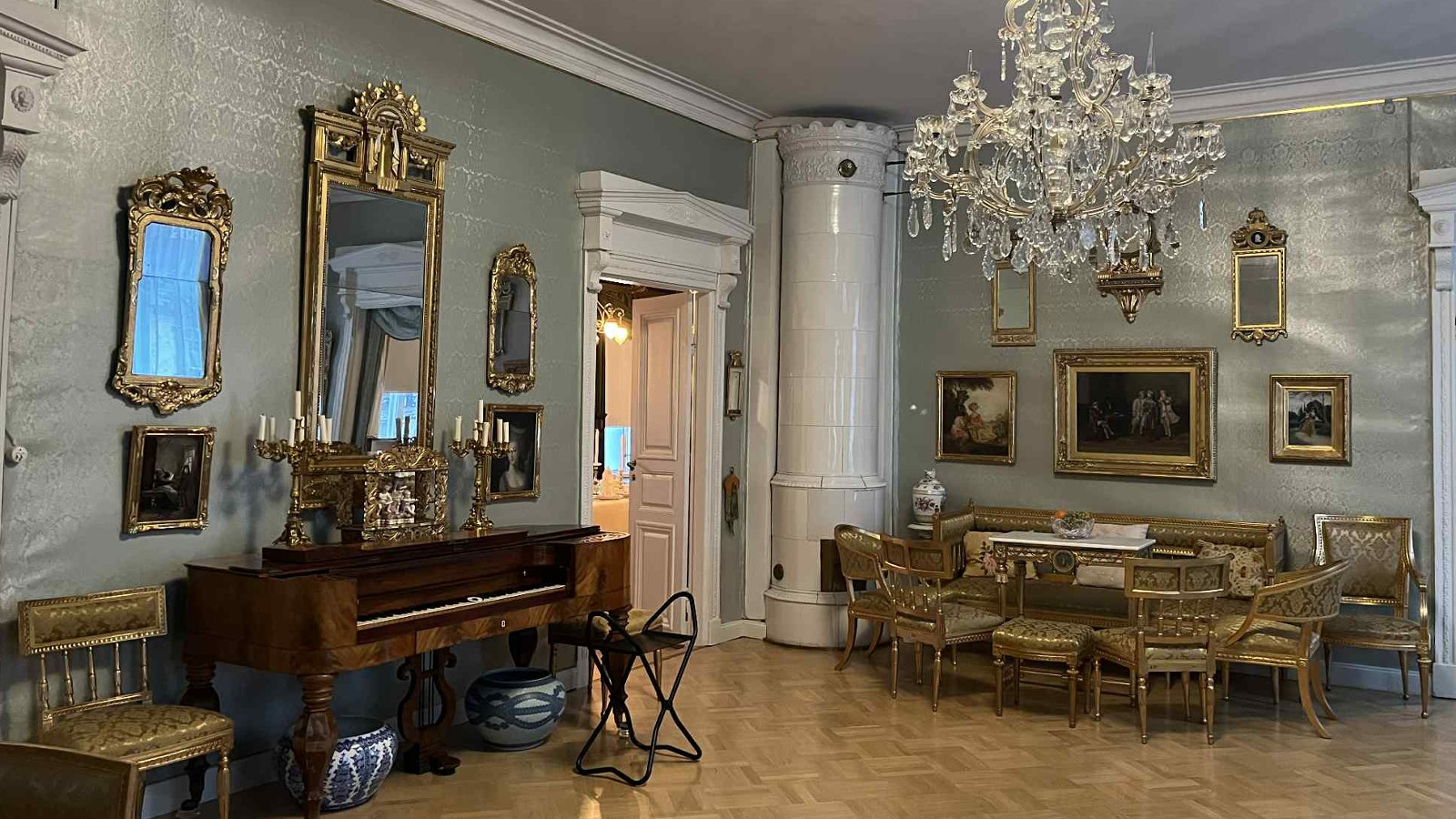 Kuvassa on huone, jossa on vanhoja huonekaluja. Vasemmalla on ruskea sembalo ja sen yläpuolella iso kultareunustettu peili. Oikealla on ruokailuryhmä.