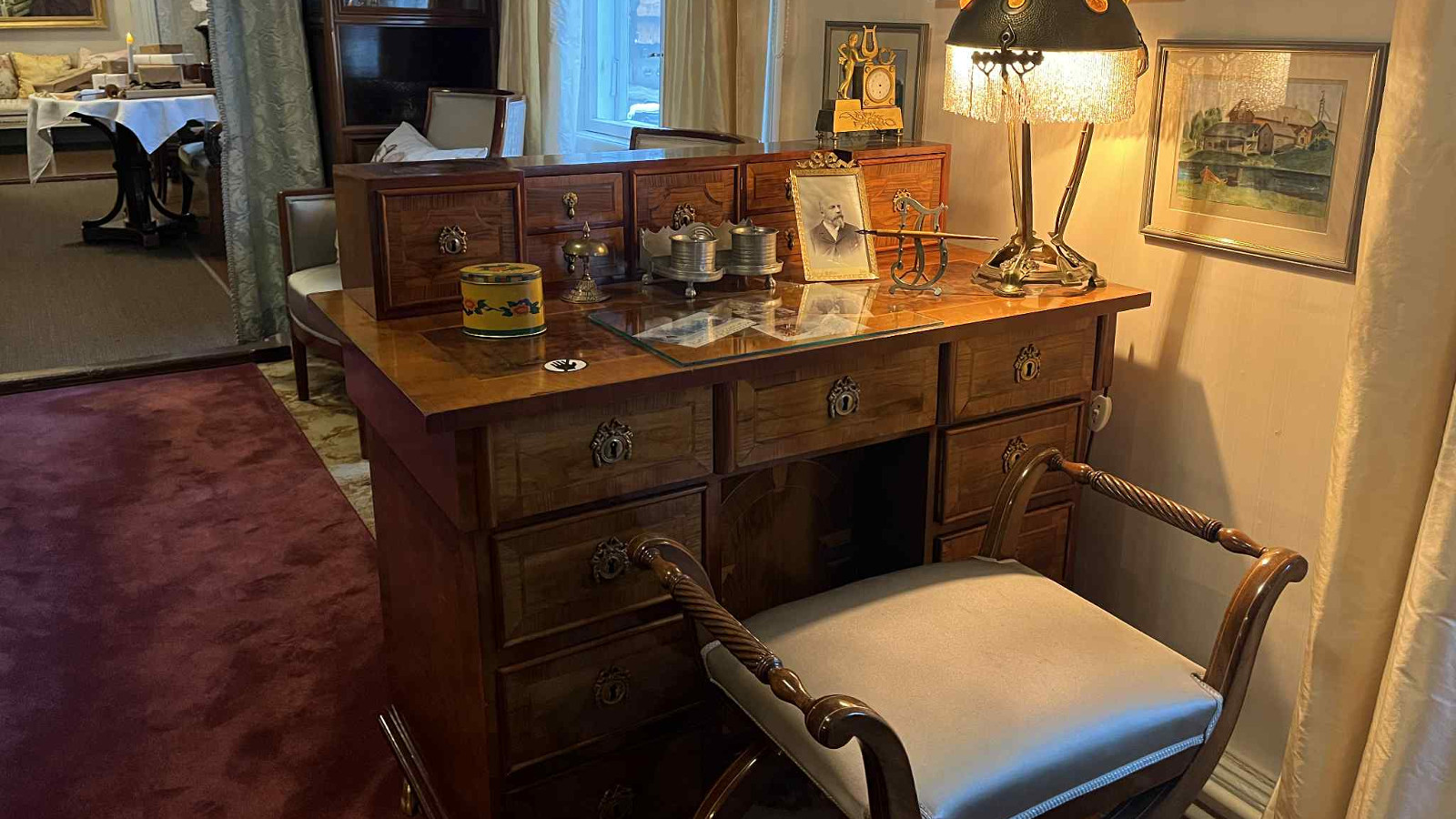 Kuvassa on huone, jossa on keskellä vanhanaikainen kirjoituspöytä, jossa on pöydällä esineitä sekä valokuva. Sen edessä on vihertävä tuoli, jossa molemmissa reunoissa kahvat.