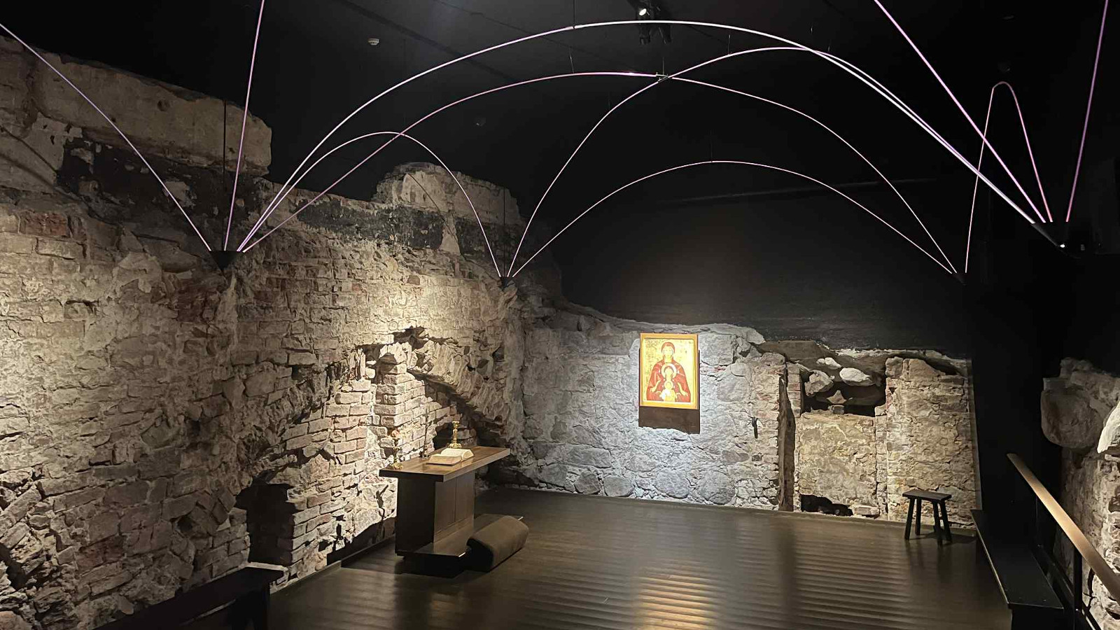 Kuvassa on tila, jossa on harmaat kiviseinät, musta lattia ja katto. Siinä on valkoiset, ylöspäin pingotetut kapeat johdot. Tilassa on teos seinällä ja veistös jalustalla.