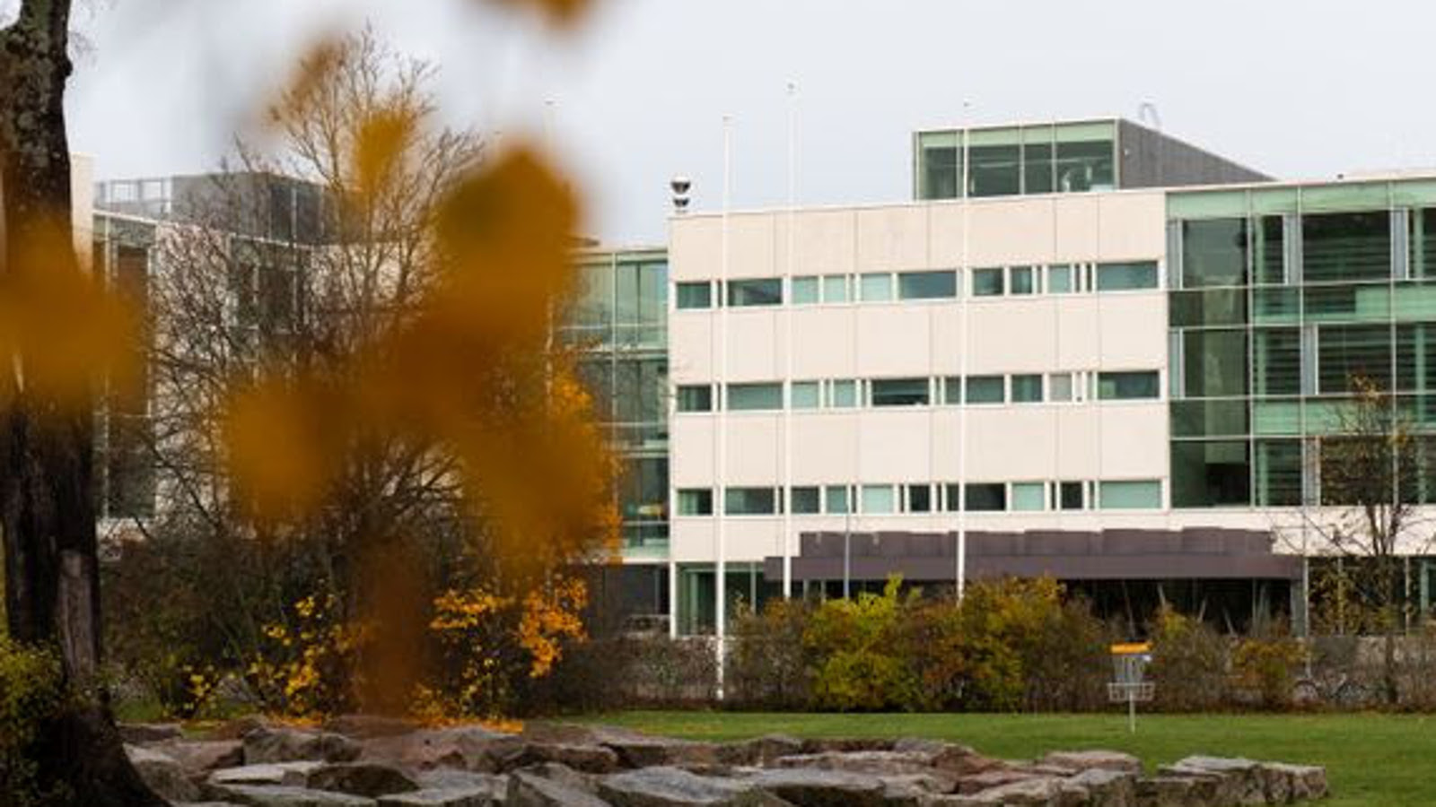Kuvassa on vasemmalla olevan lähes lehdettomän puun takana oleva rakennus, joka on L.n muotoinen ja jonka oikea osa on vaalea. Alla näkyy litteähköjä kiviä ja pihapensaita