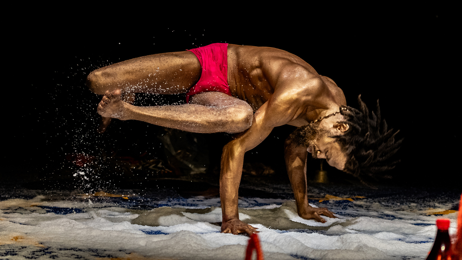 Kuvassa mies, jolla on pronssiseksi maalattu iho, ja hän käsien päällä ja muu keho vinottain vaaka-asennossa ylhäällä. Hänellä on punaiset alushousut. Tausta on musta ja lattialla on valkoista vaahtoa.