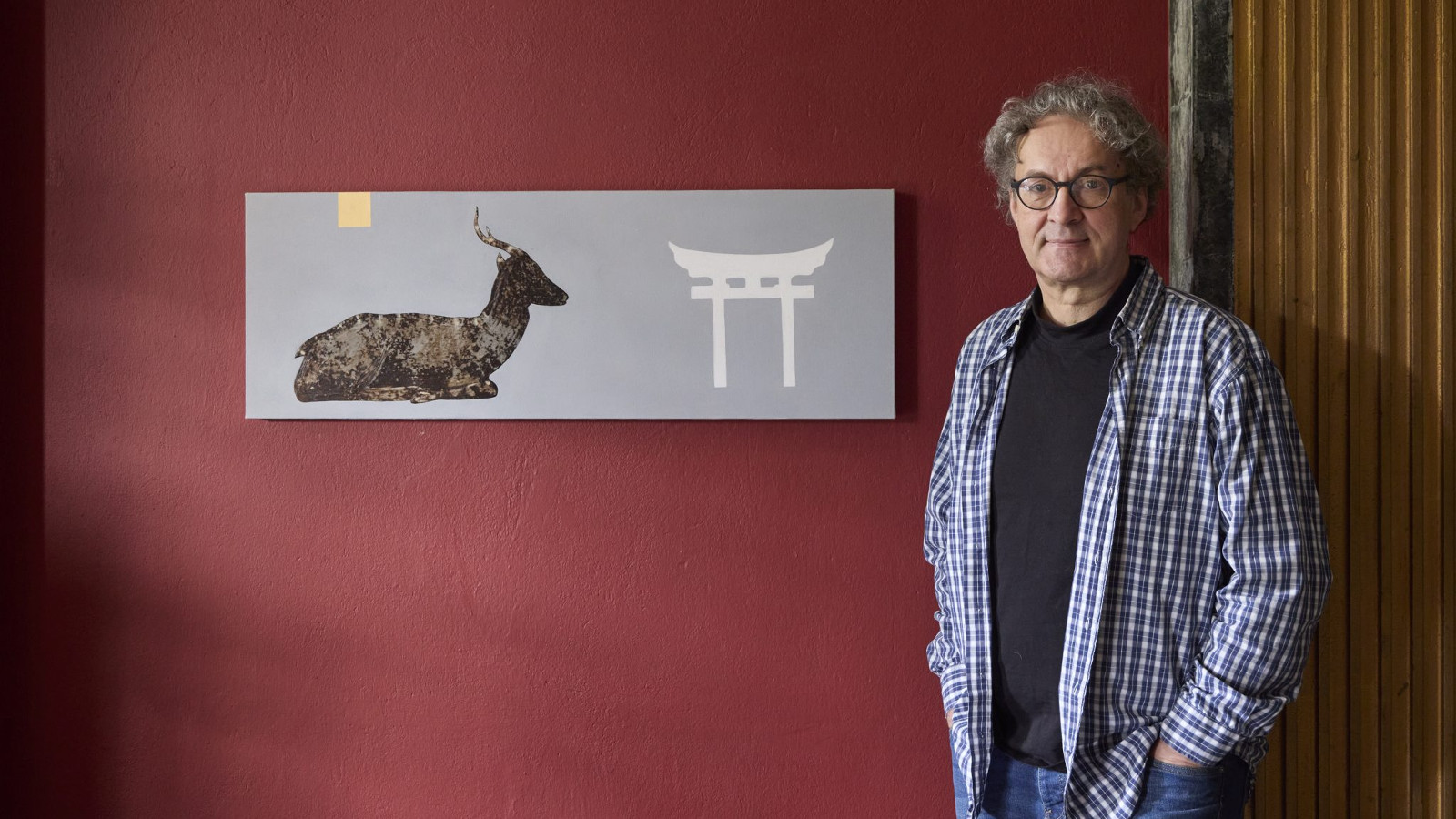 Kuvassa on Osmo Rauhala sini-valkoruutuisessa paidassa seisomassa oikealla. Burgundin värisellä seinällä on suorakaiteen muotoinen teos, jossa on vasemmalla tumma kauris makaamassa sivuttain ja japanilainen pyhä portti.